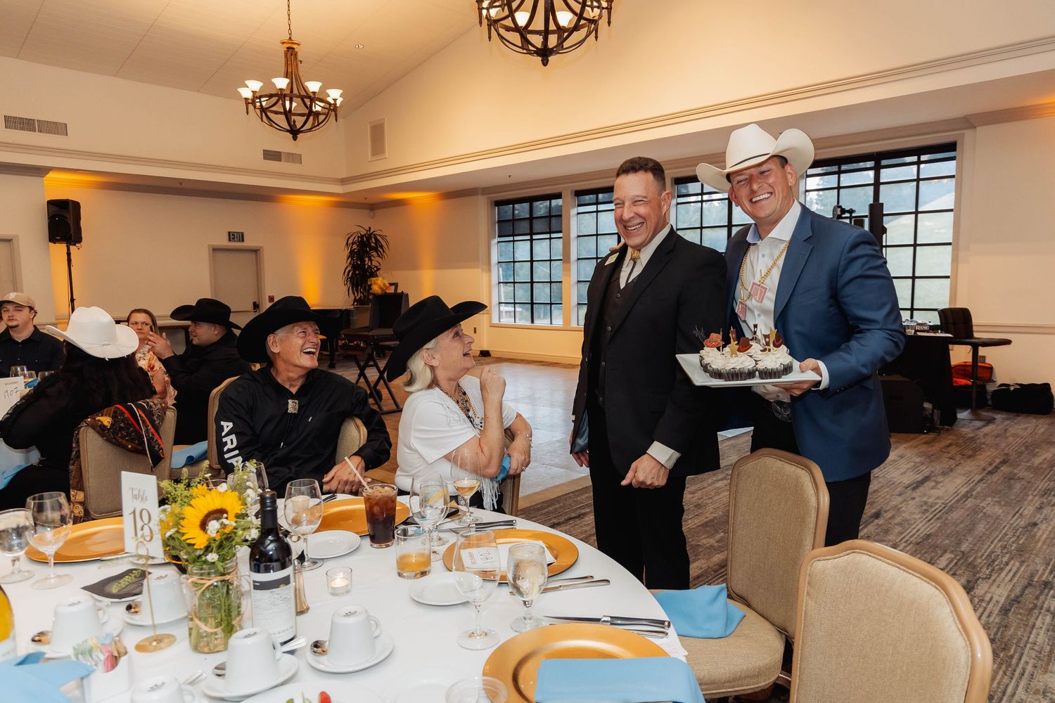 Two men in suits and cowboy hats present a birthday cake to seated guests at an elegant country club dinner.
