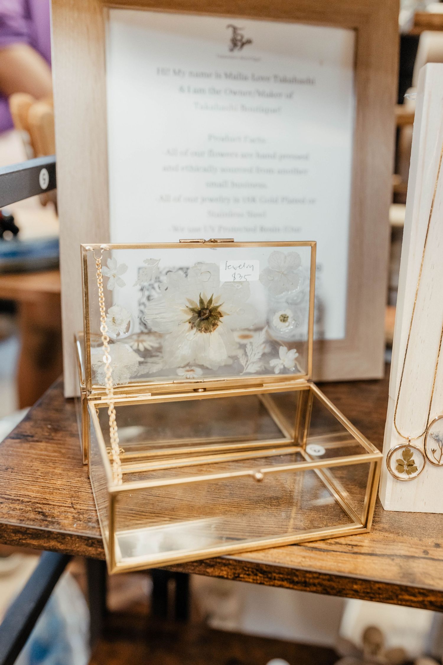 Gold-framed glass display box with delicate pressed flower on wooden surface.