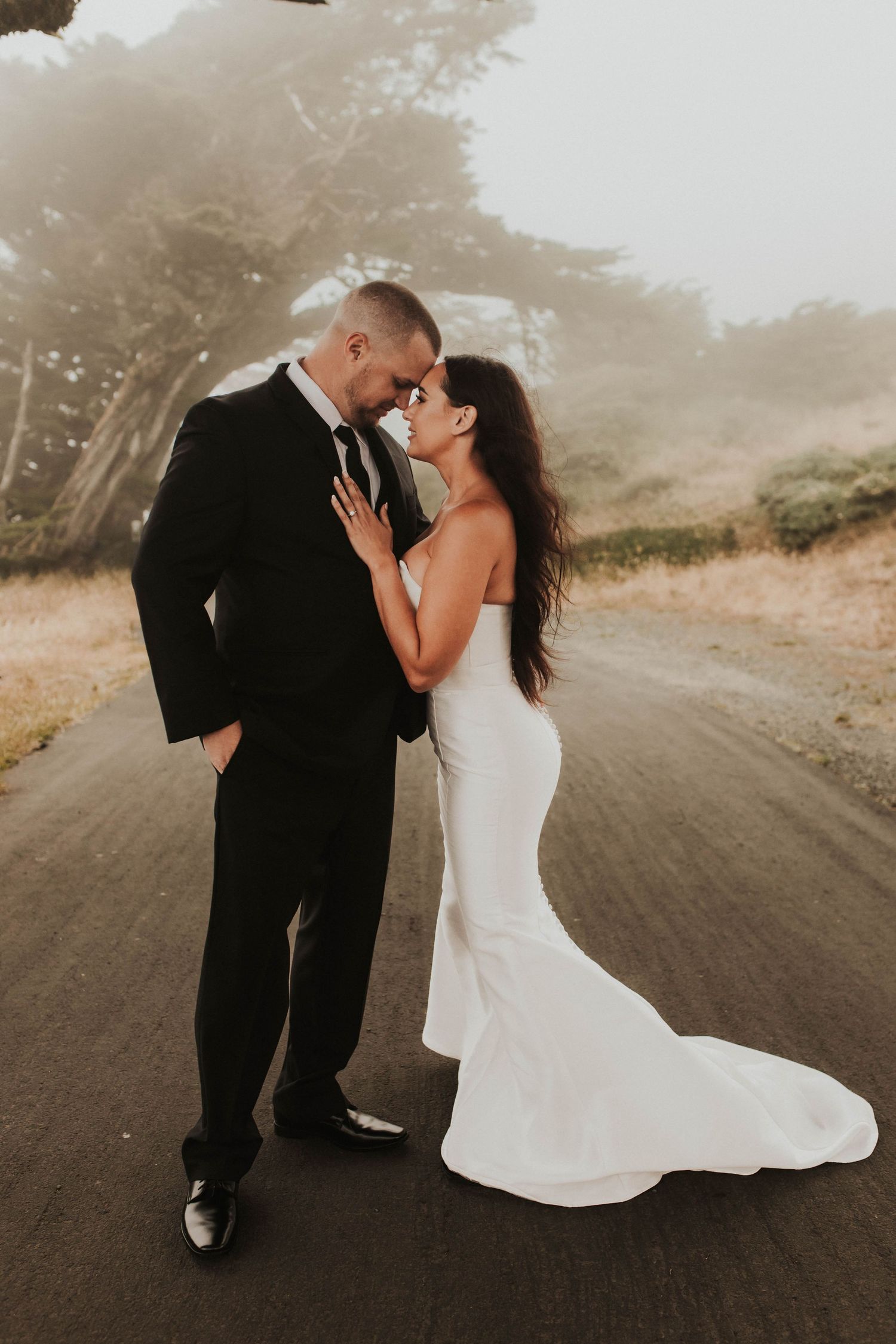 A romantic wedding couple embraces on a foggy dirt road wearing classic black suit and white mermaid gown.