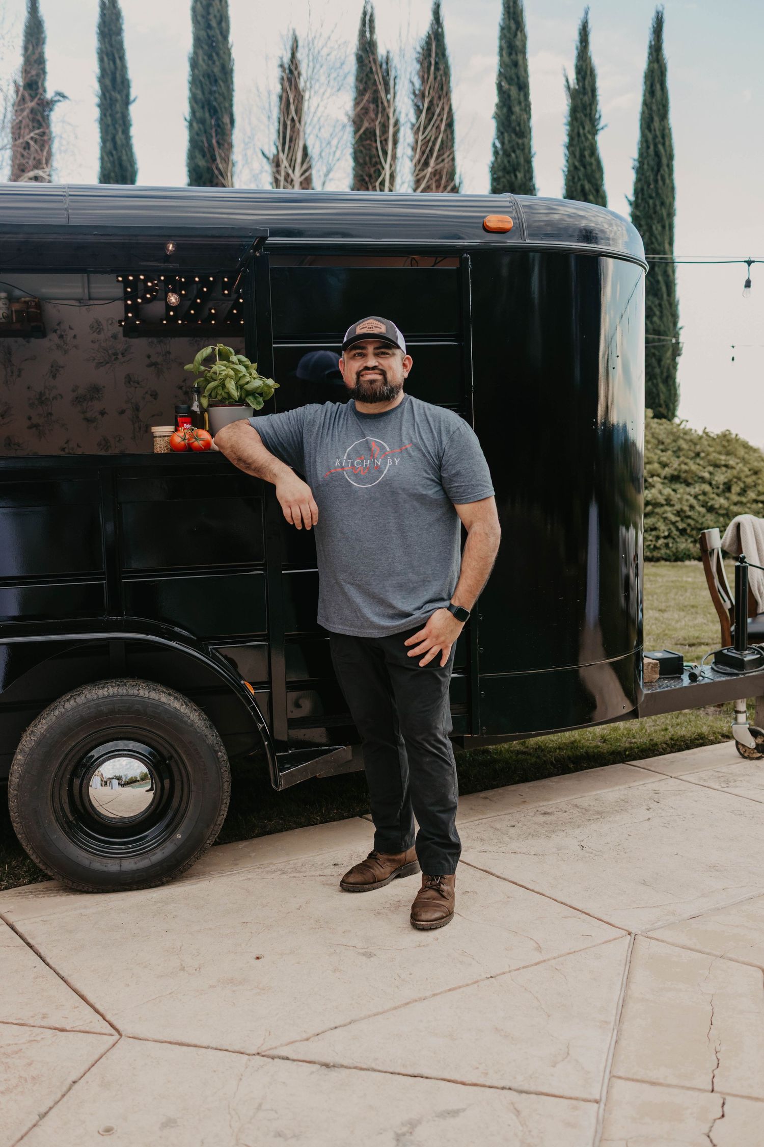Black food truck parked on concrete with cypress trees in the background.