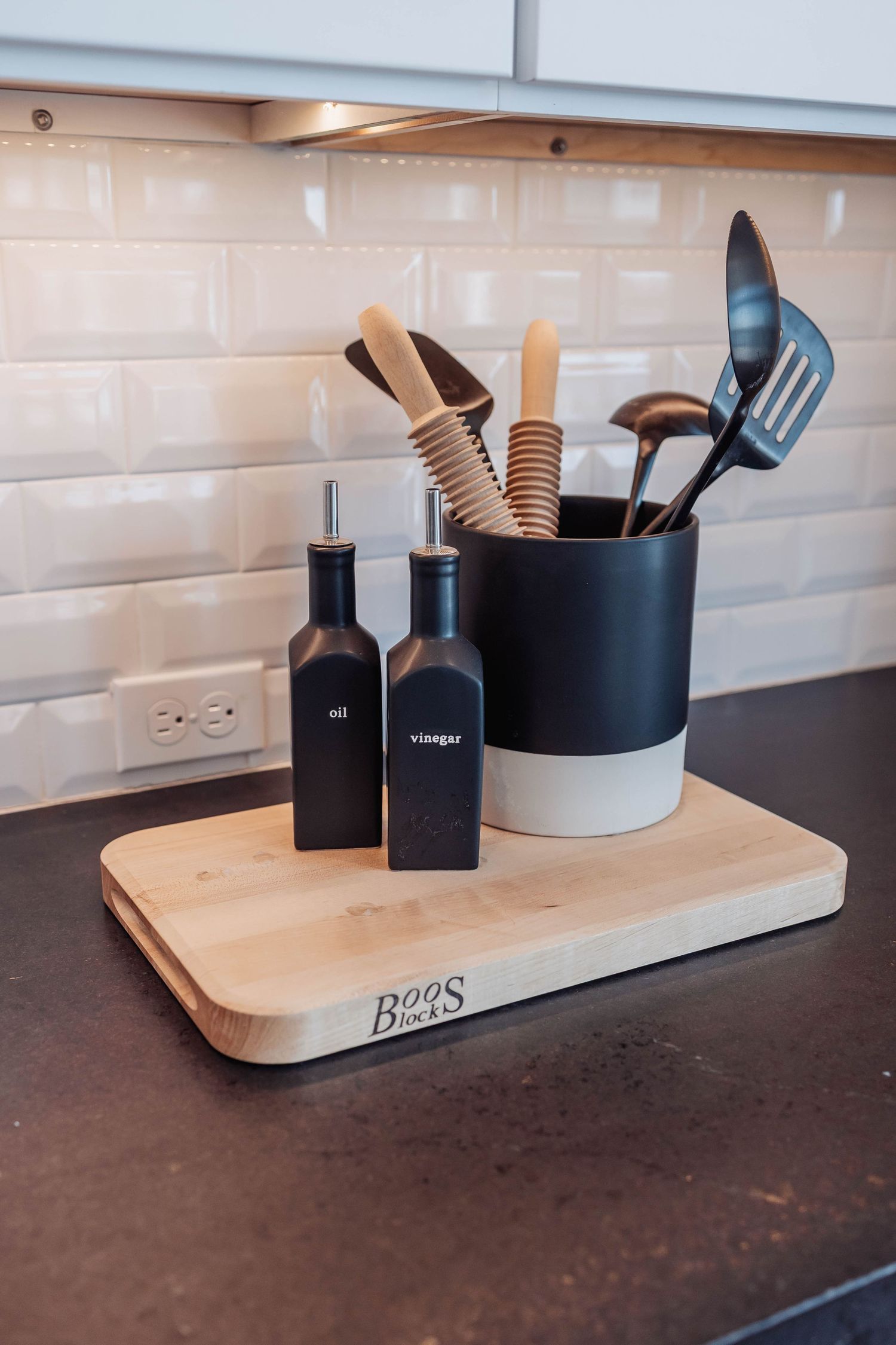 Kitchen counter display with wooden utensils in a black and white holder alongside dark bottles on a cutting board.