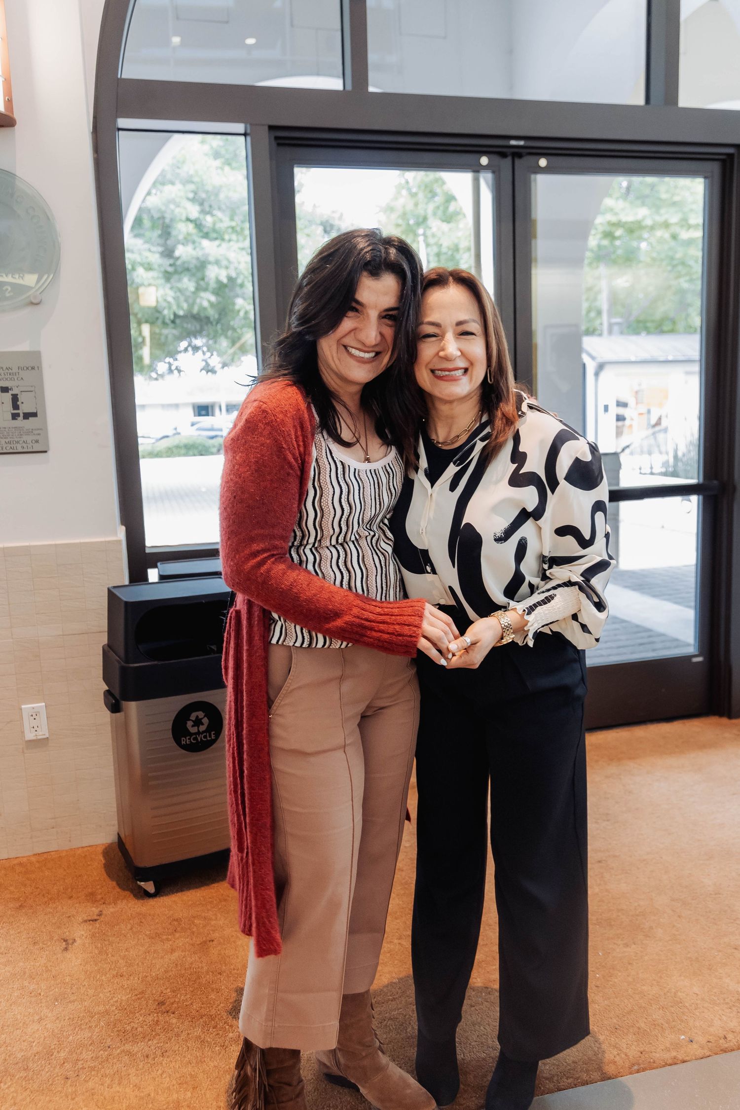 Two women smiling and embracing inside a modern building entrance, one wearing a red cardigan and the other a black and white blouse.