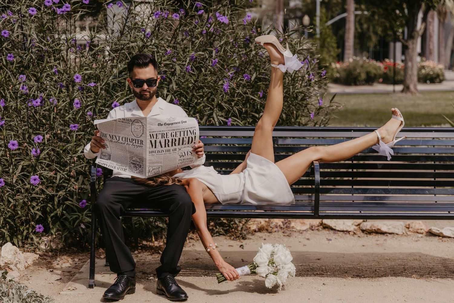 Playful engagement photo with couple reading newspaper on park bench with legs in air.