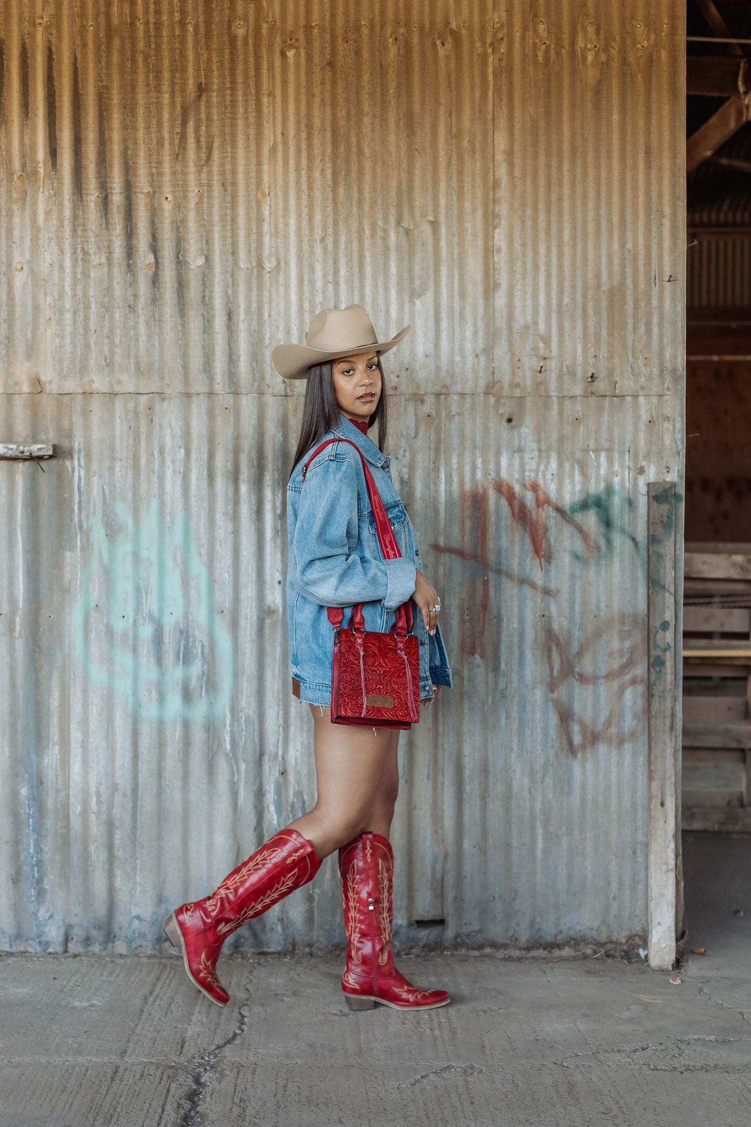 Western-inspired outfit featuring red boots, denim jacket and matching red handbag against metal wall.