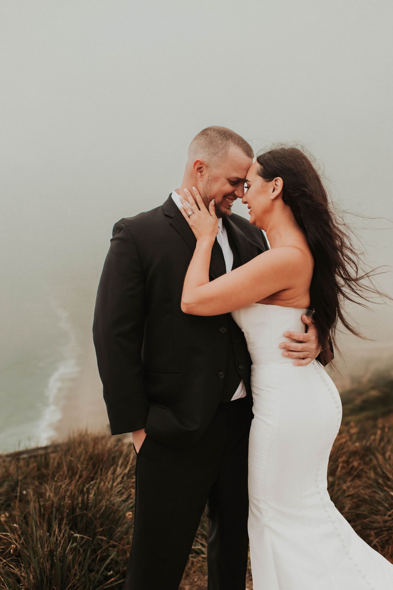A joyful wedding couple shares a laugh while embracing on a coastal cliff.