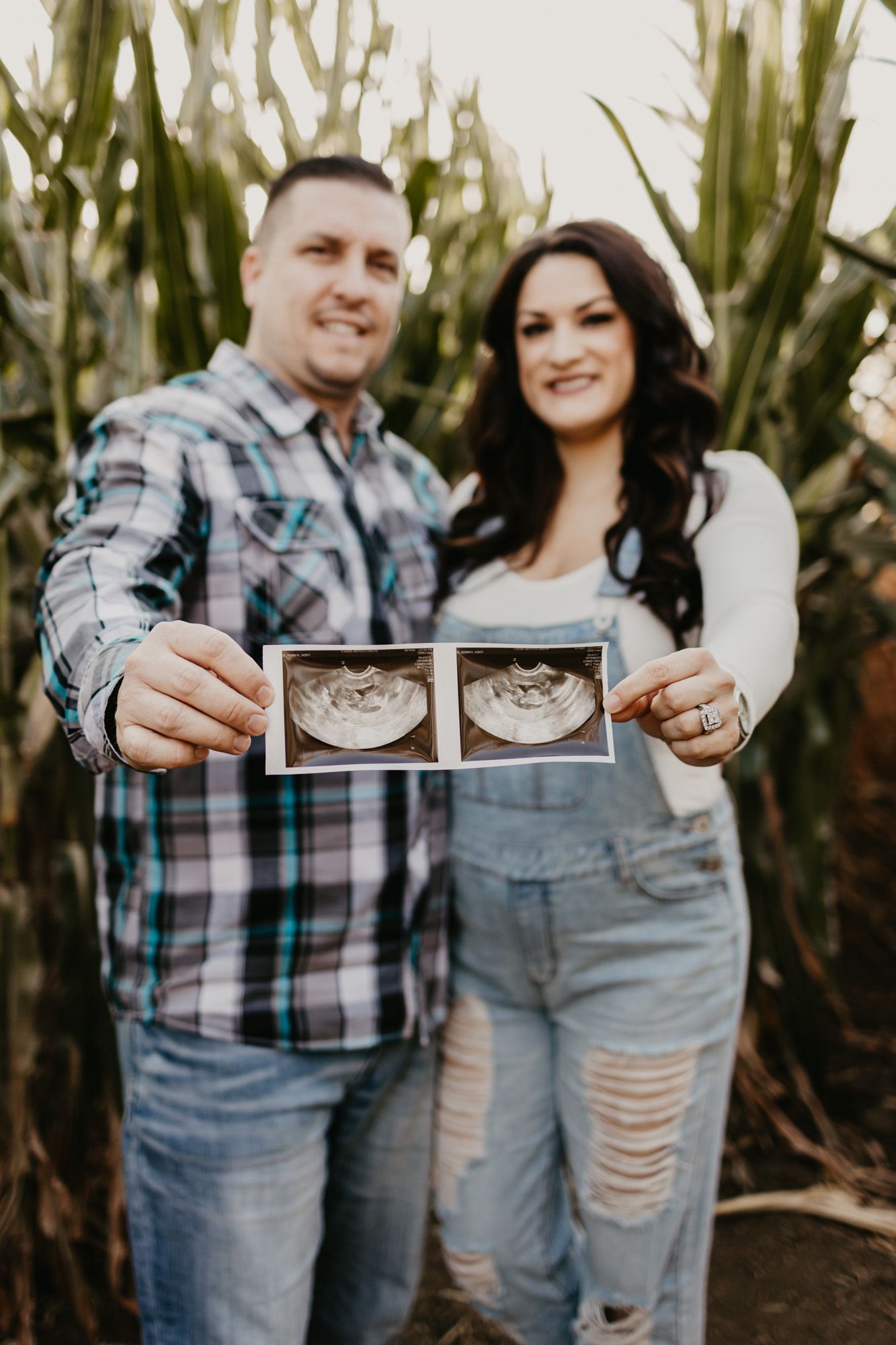 Couple smiling while displaying sonogram pictures against tall corn stalks in the background.