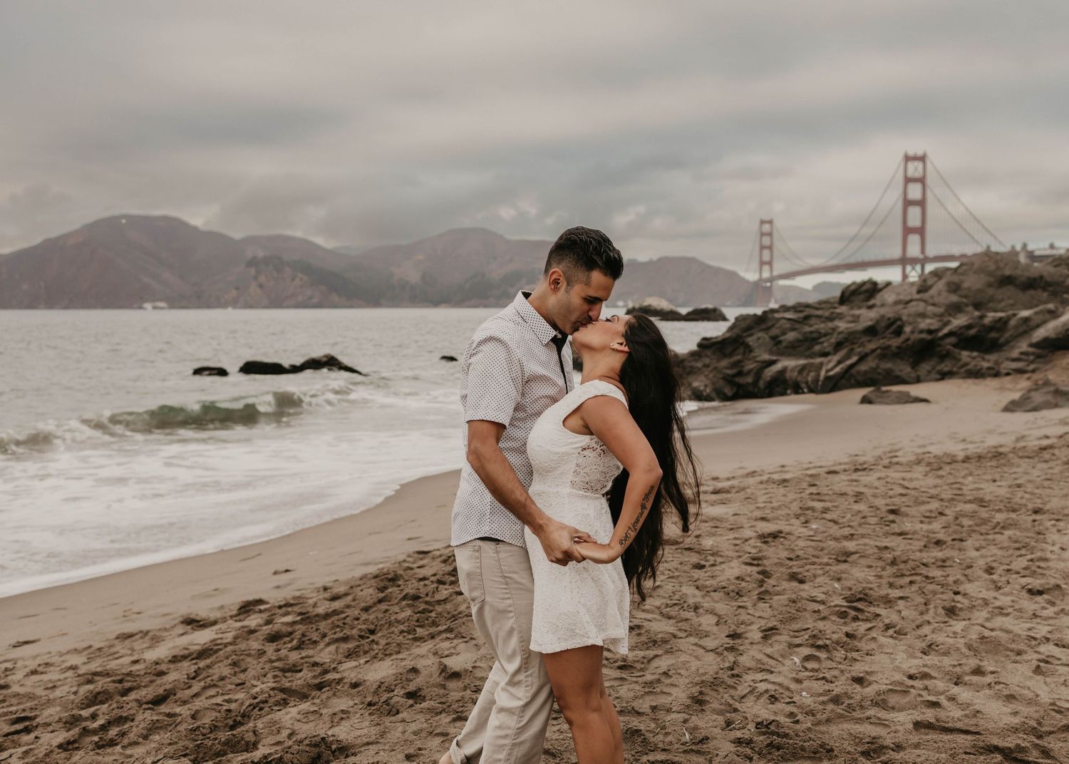 Couple embracing on sandy beach with San Francisco's Golden Gate Bridge in distance.