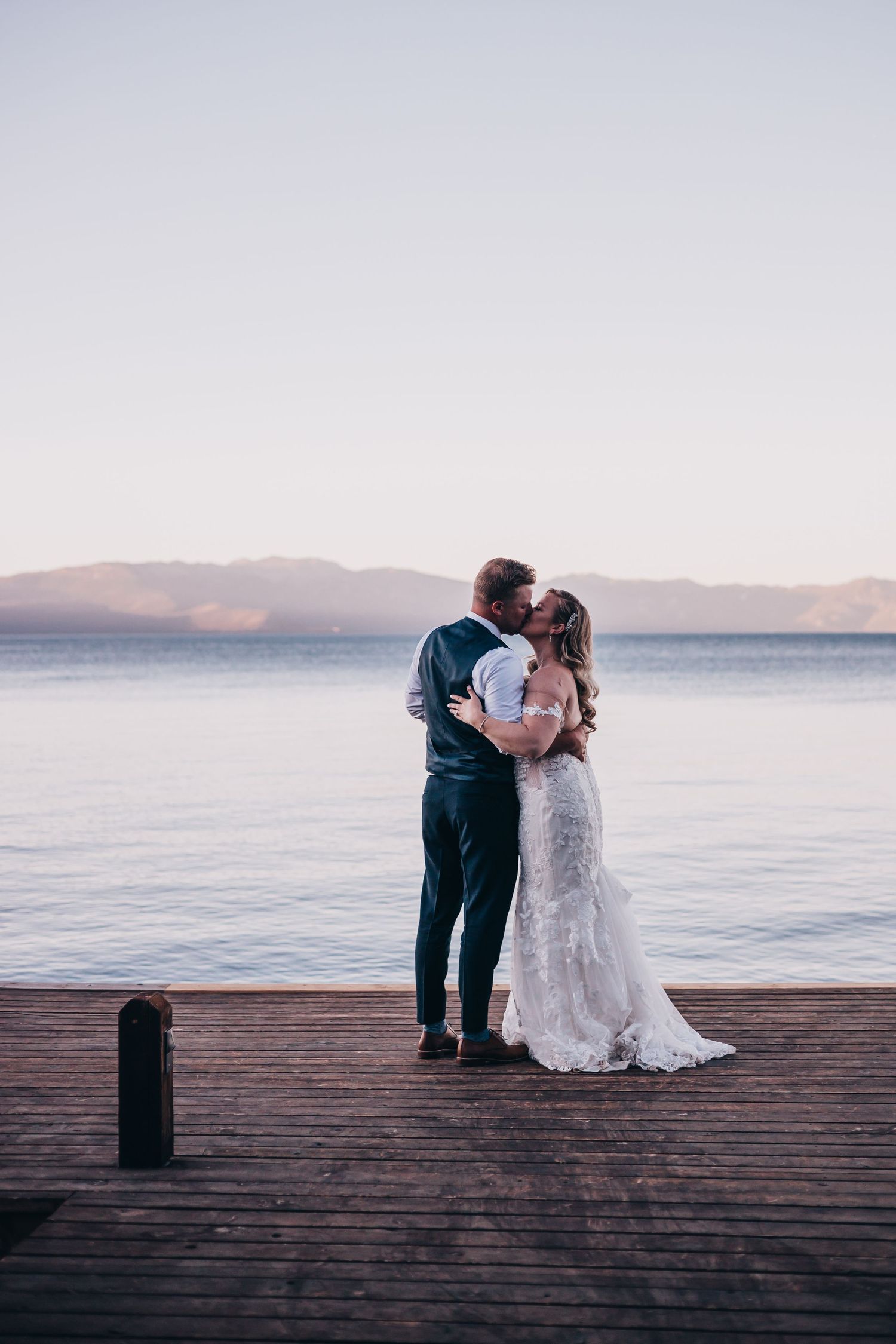 Wedding couple embracing on wooden pier overlooking serene lake at sunset.