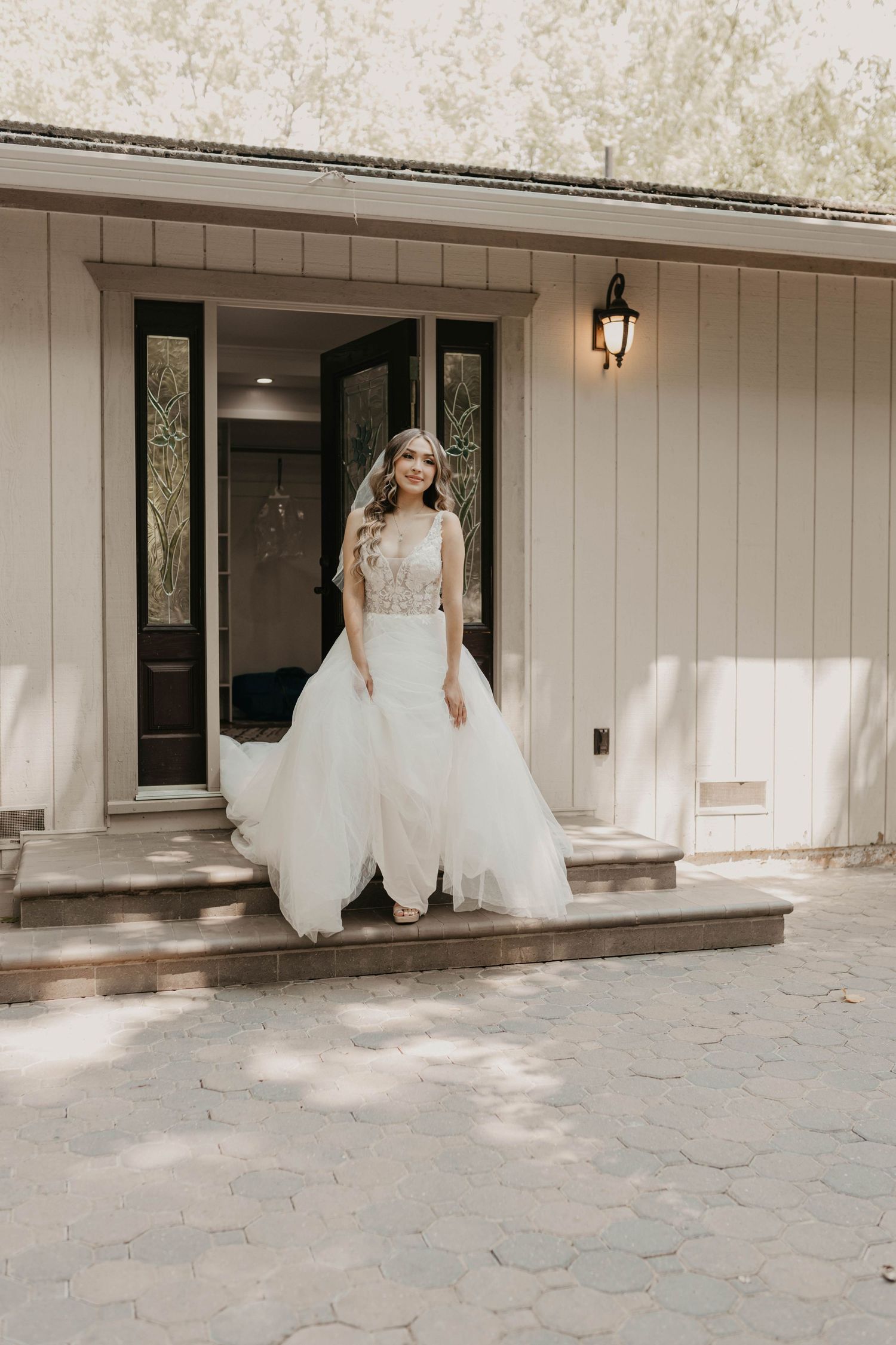 A bride in a flowing white wedding gown twirls and dances on a stone patio outside a light-colored building.