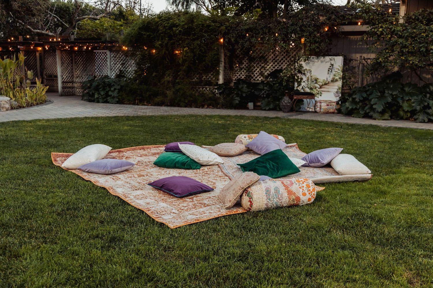 Colorful throw pillows arranged on a patterned outdoor rug on green grass, with string lights and garden backdrop.