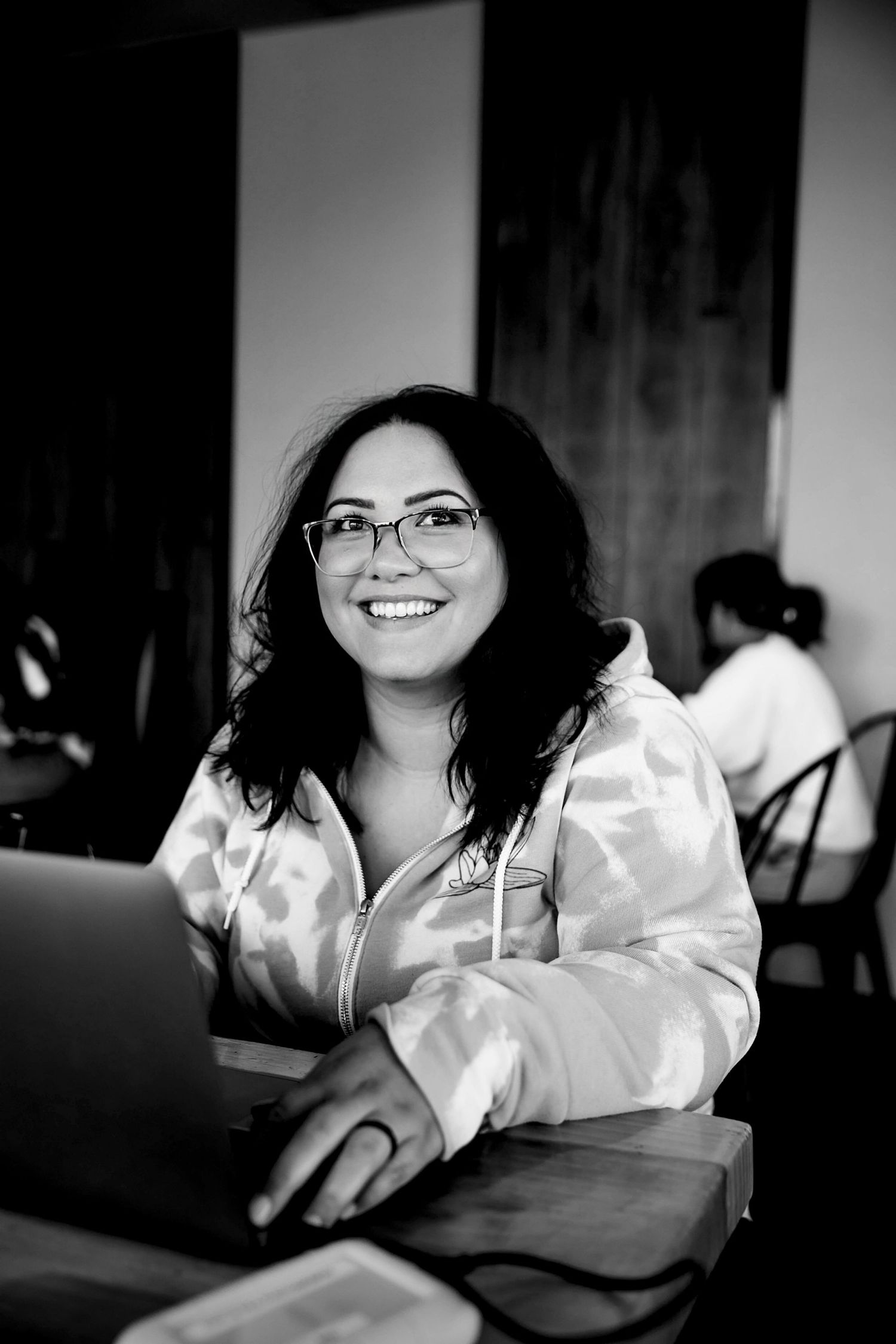 Smiling woman with glasses working on laptop at cafe table in black and white photo.