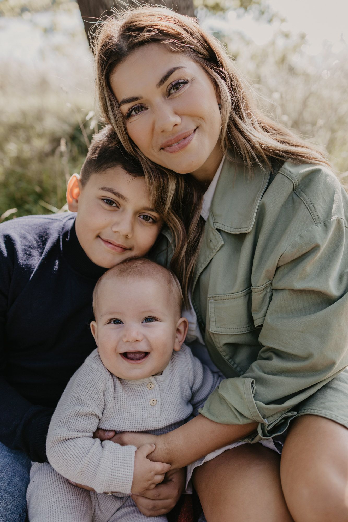 Heartwarming outdoor family portrait showing warm embraces between young siblings and parent in natural lighting.