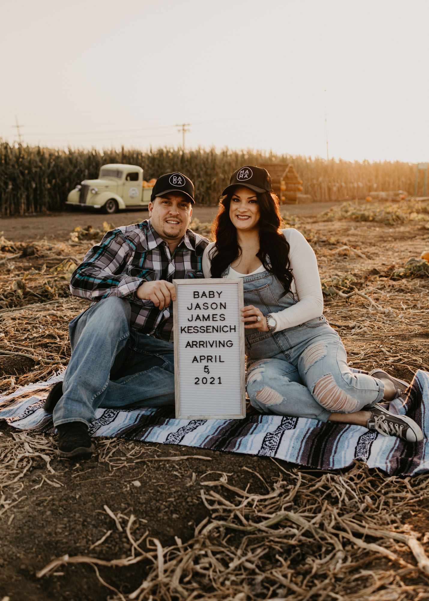 Couple announces pregnancy with letterboard sign while sitting on blanket in rural setting.