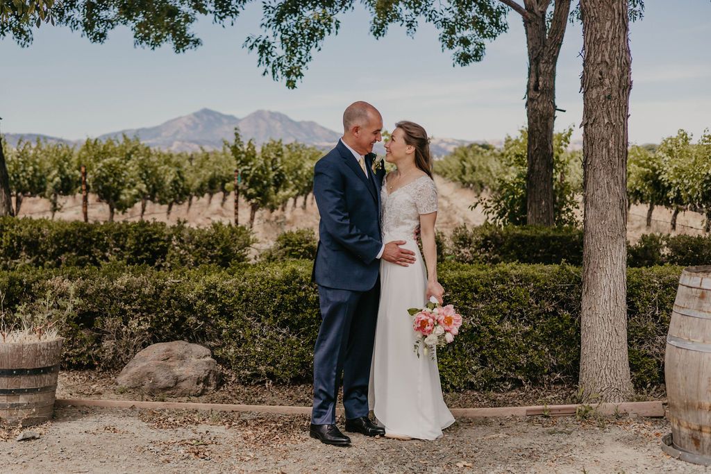 Wedding couple poses in front of vineyard rows with mountain backdrop.