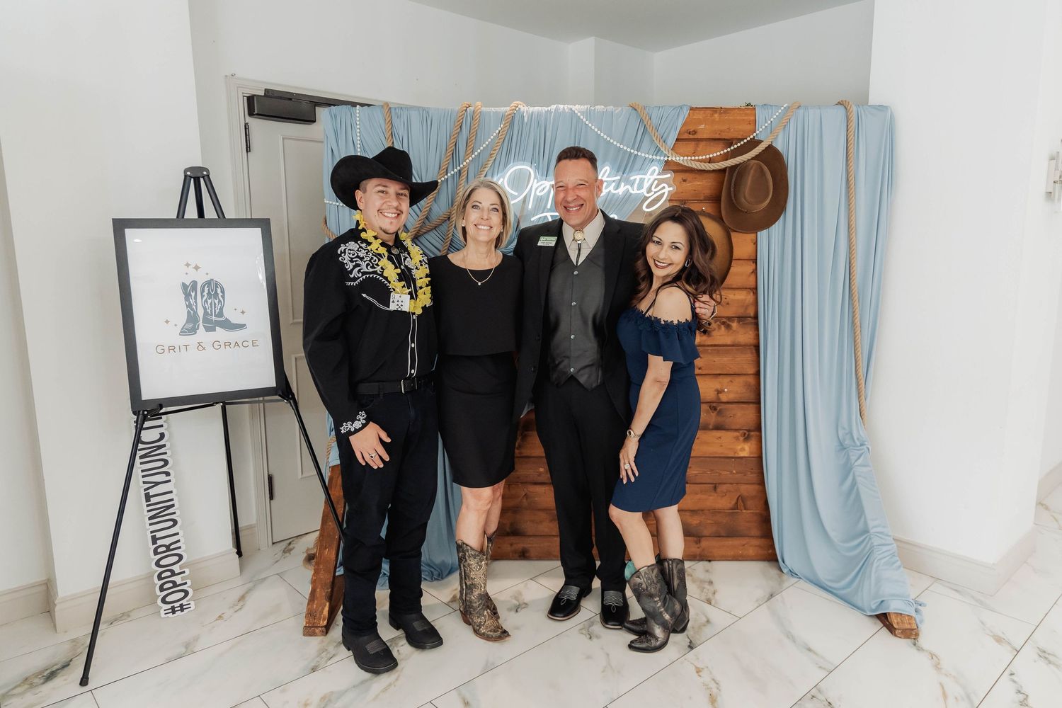 Four people posing together at a western-themed event with cowboy hats and rustic wood backdrop.