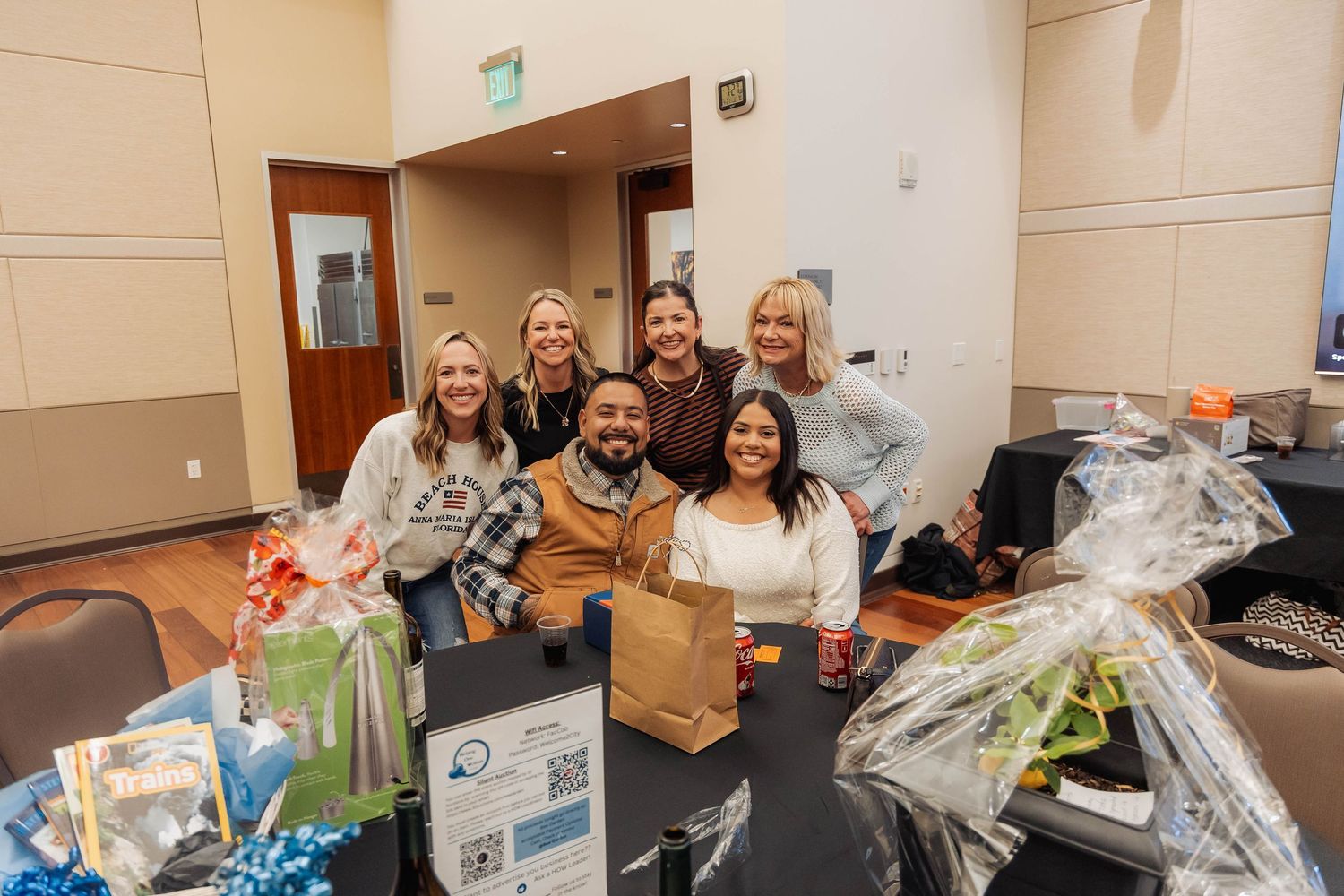 Group of six smiling colleagues posing together at a table with gift baskets at an indoor corporate event.