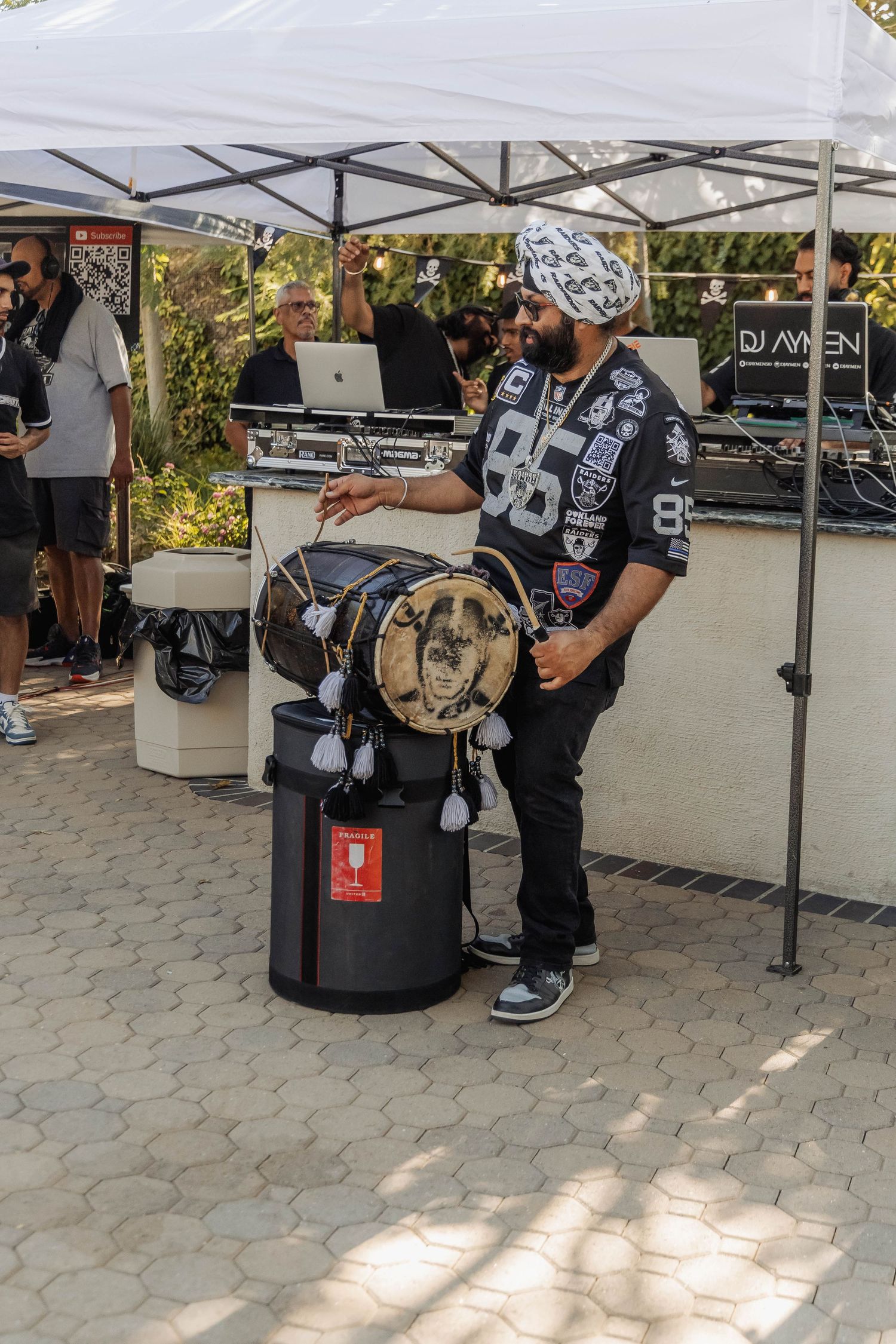 Man playing a decorative drum at an outdoor music event with DJ setup and crowd under a white tent.