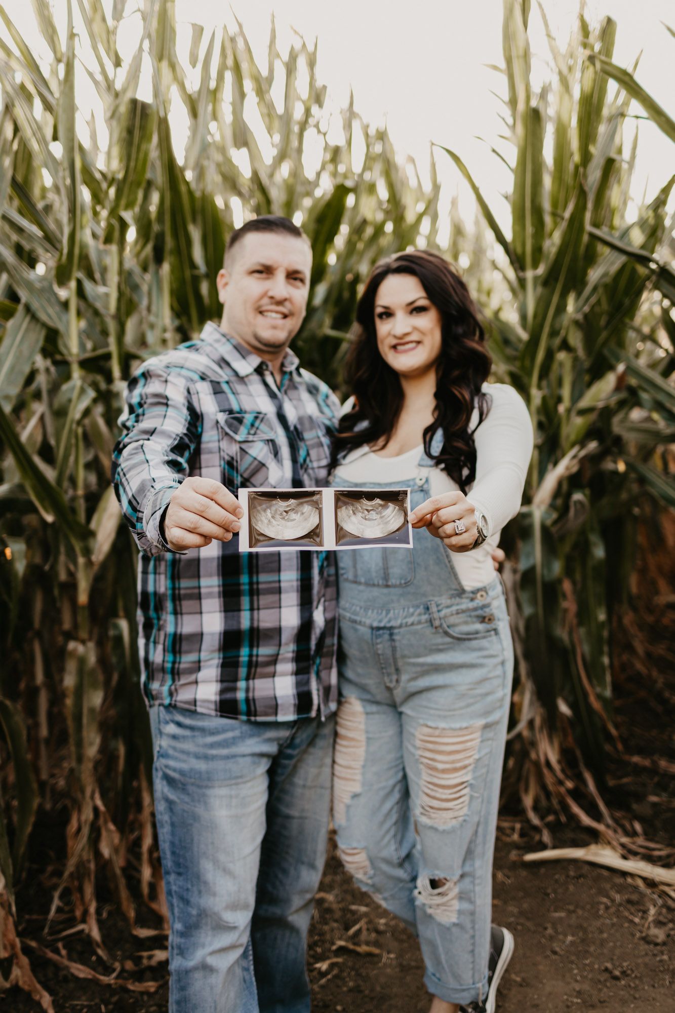 Couple holding ultrasound photos while standing in front of a cornfield during pregnancy announcement photoshoot.