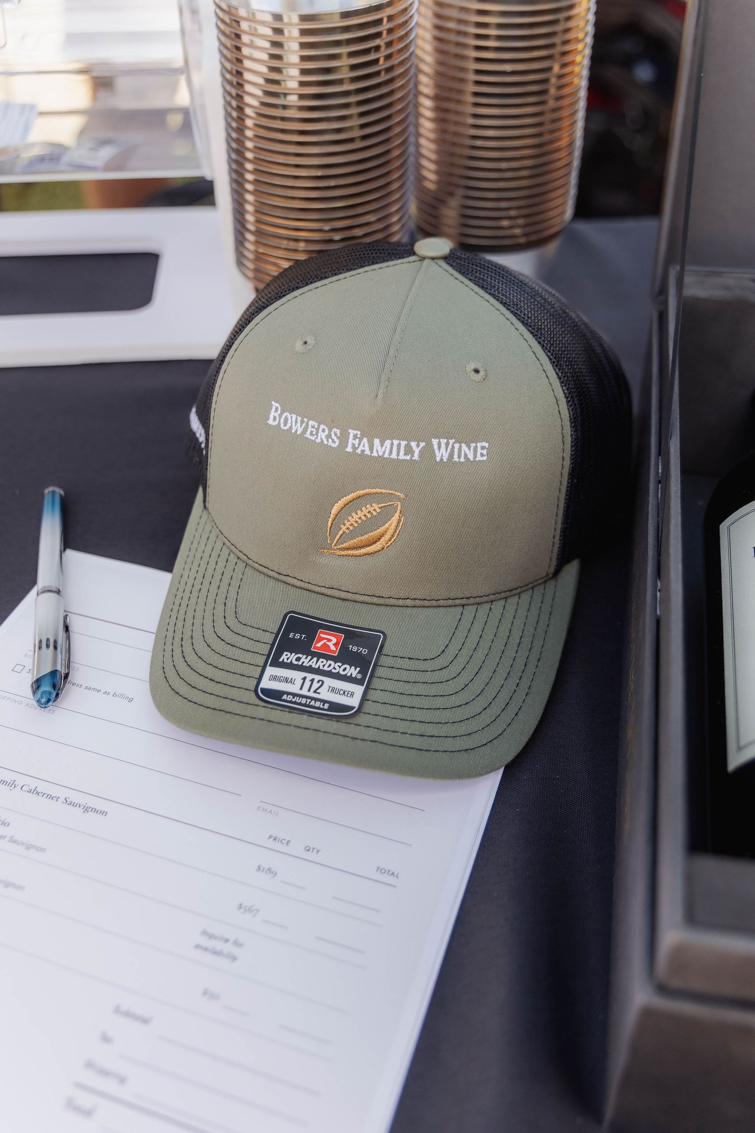 Olive green and black Richardson 112 trucker hat with Bowers Family Wine embroidery on a clipboard at an outdoor event.