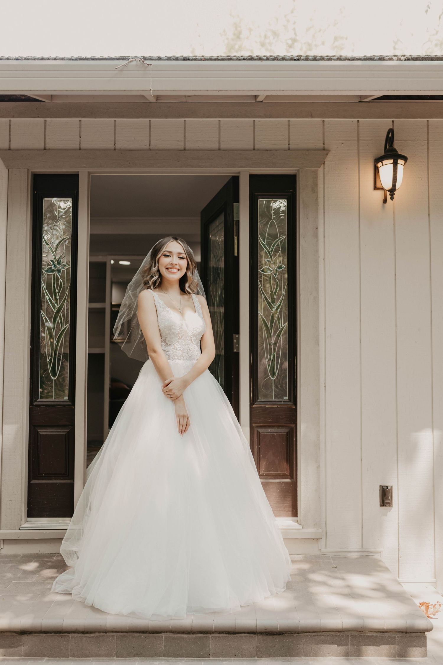 A bride in a flowing white ballgown wedding dress stands elegantly on a porch with classic wooden doors and lantern.
