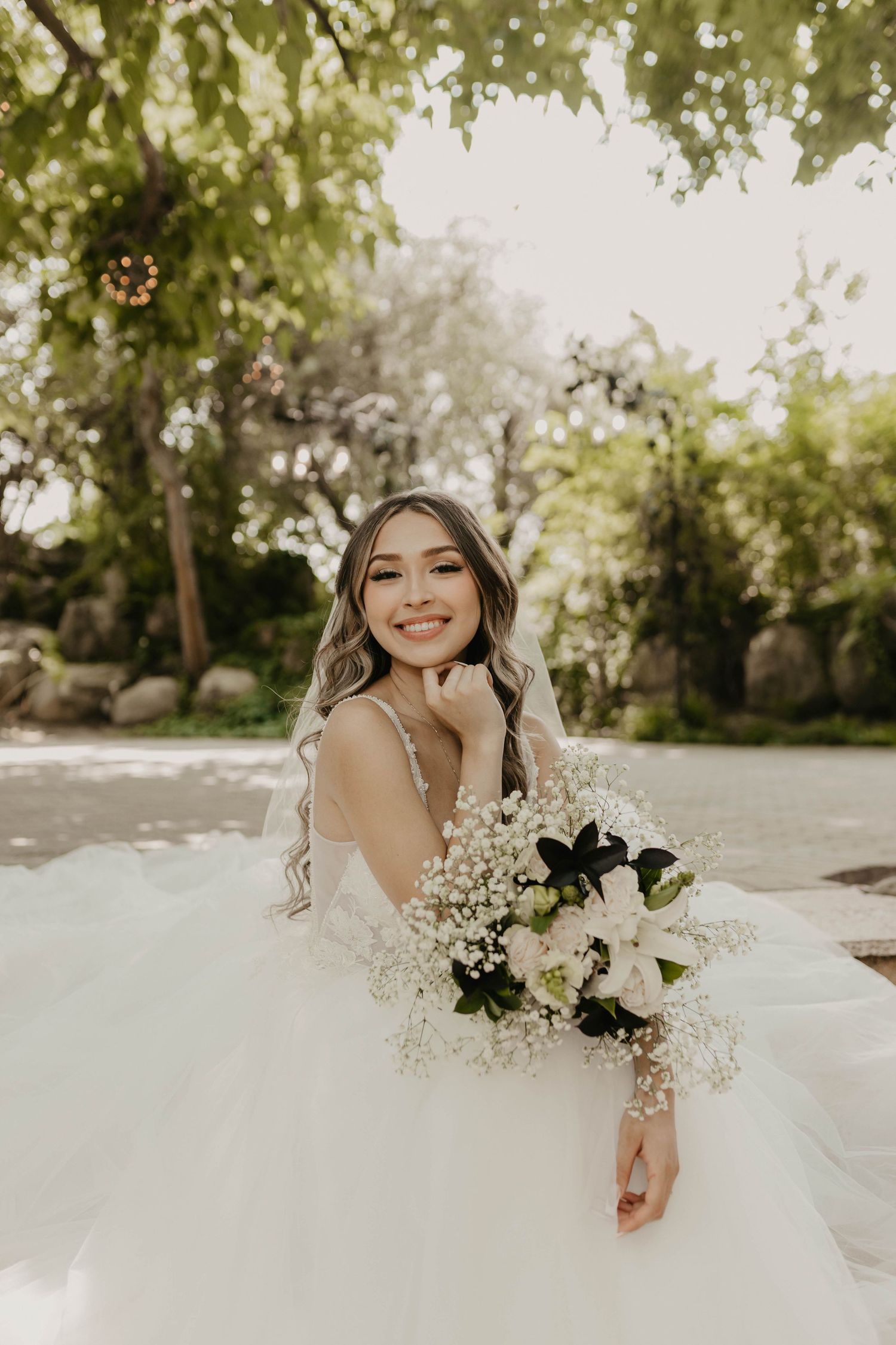 A bride in a white tulle gown holds a bouquet of white lilies and baby's breath in a sunlit garden.