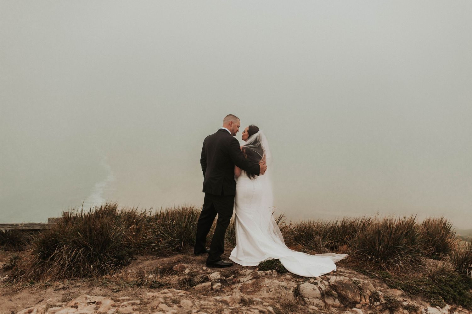 A couple shares an intimate moment in wedding attire on a misty hilltop surrounded by coastal grass.
