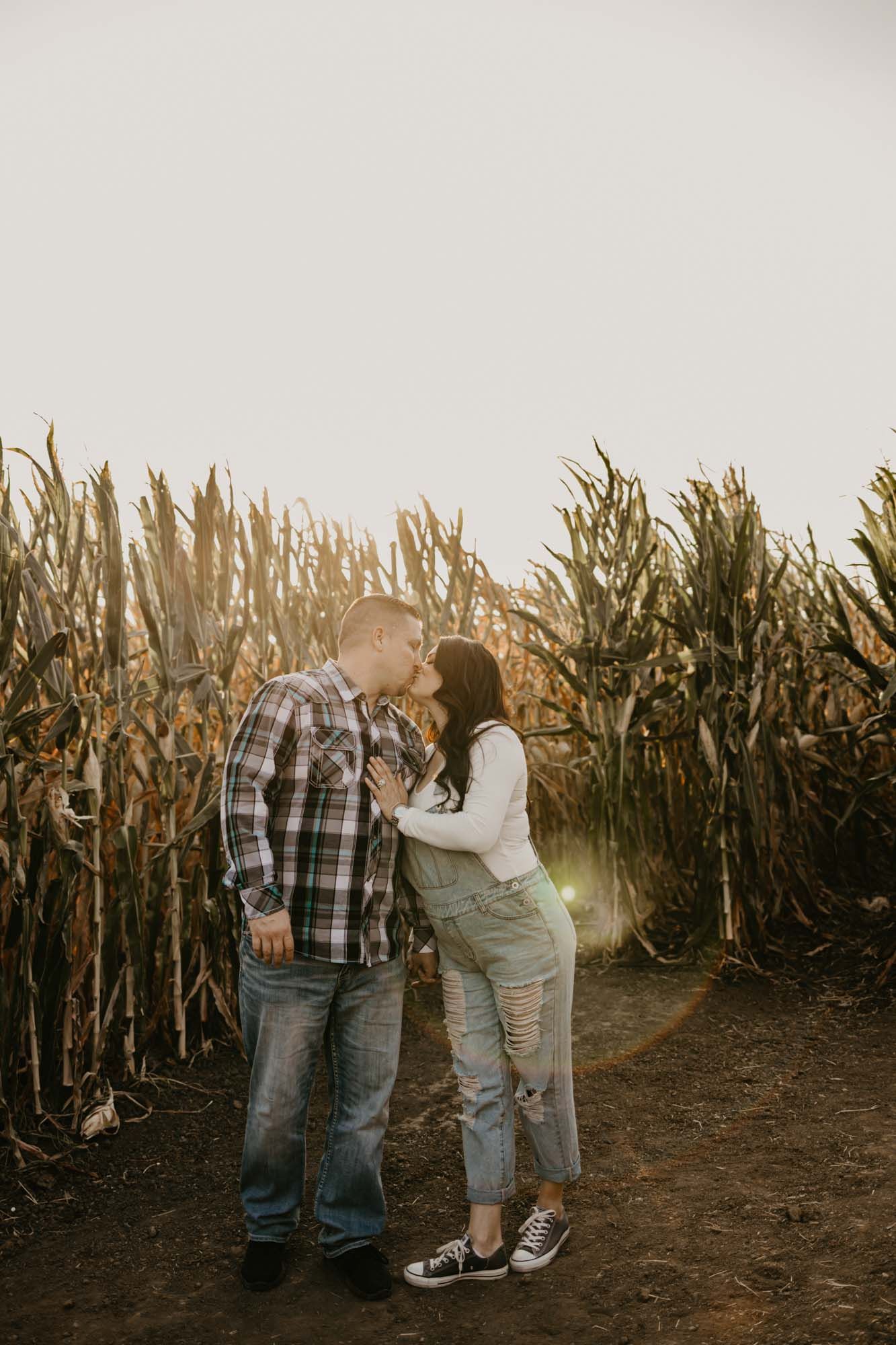 A romantic moment captured between a couple in overalls and plaid standing amid cornfields.