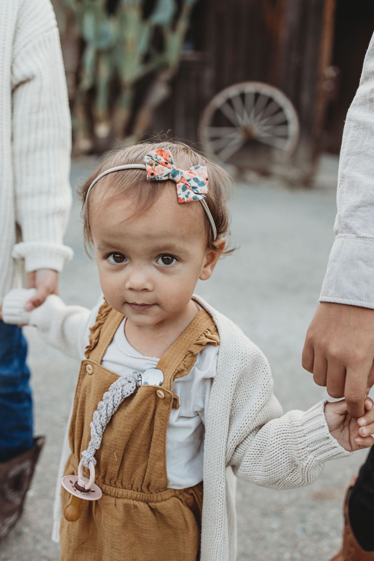 Toddler in brown overalls and floral headband holding hands with family.