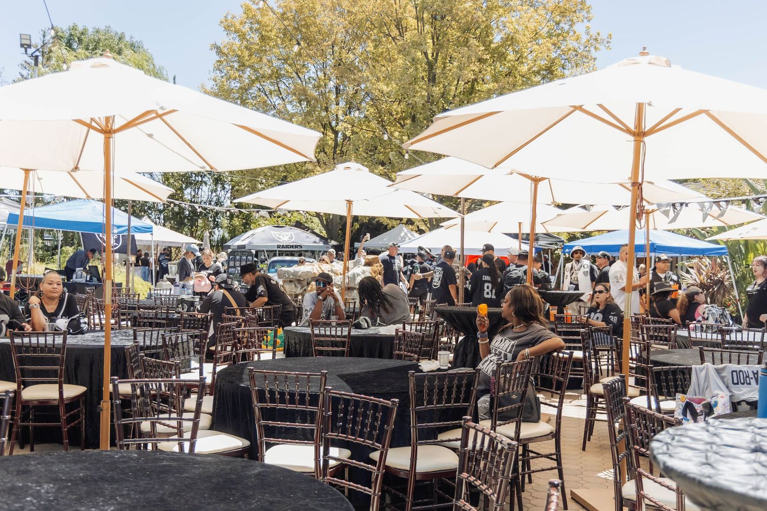Outdoor dining area with white umbrellas, dark chairs and tables, filled with guests enjoying a sunny day event.