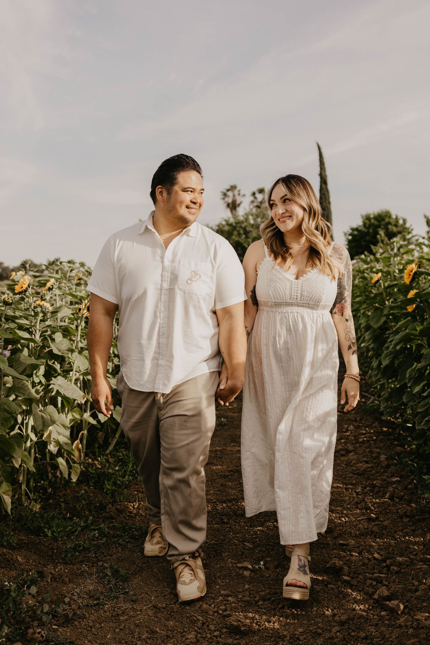 A couple walking hand in hand through a garden path wearing coordinated white and beige outfits.