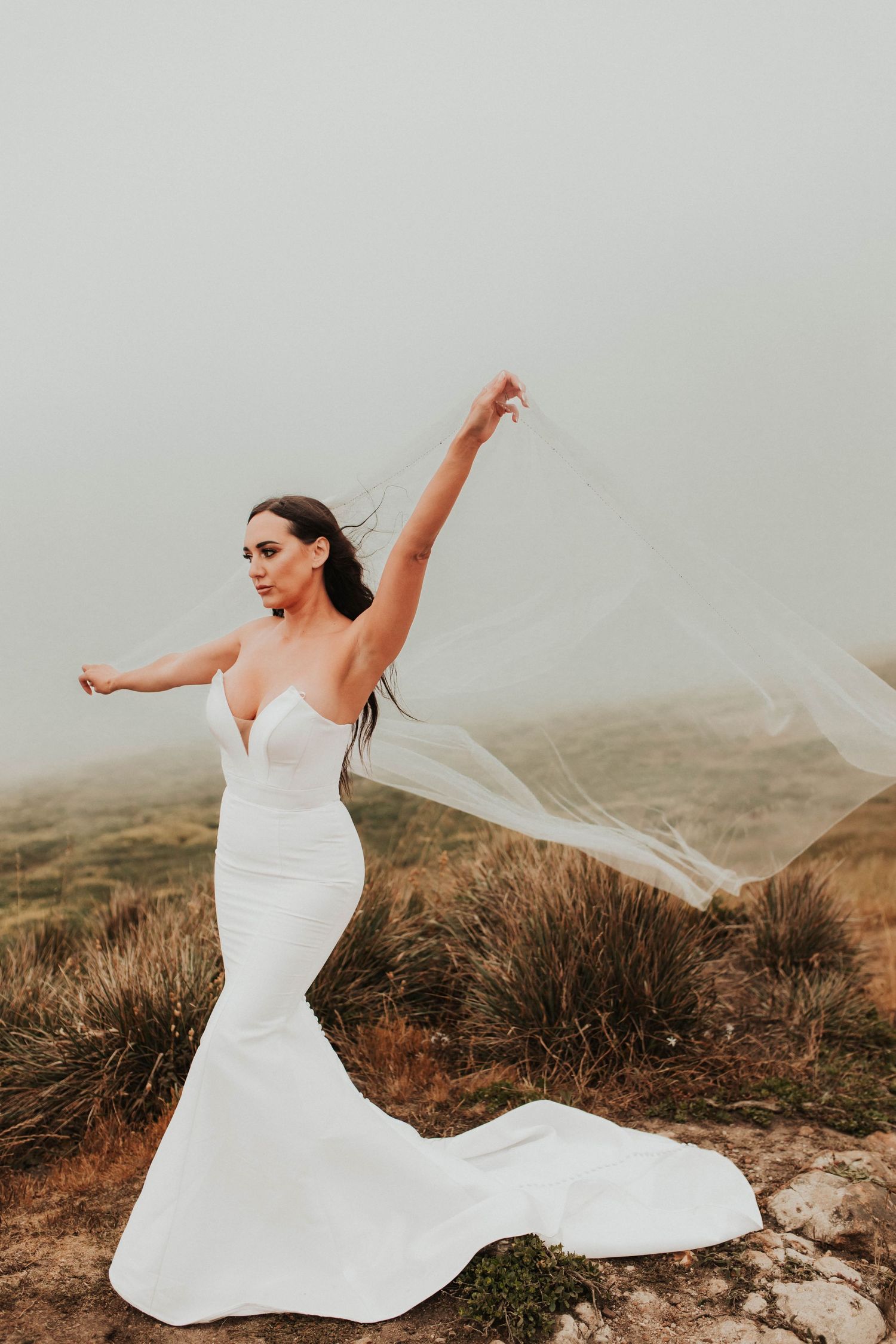 A bride in a fitted white dress poses dramatically with flowing veil on a cliff.