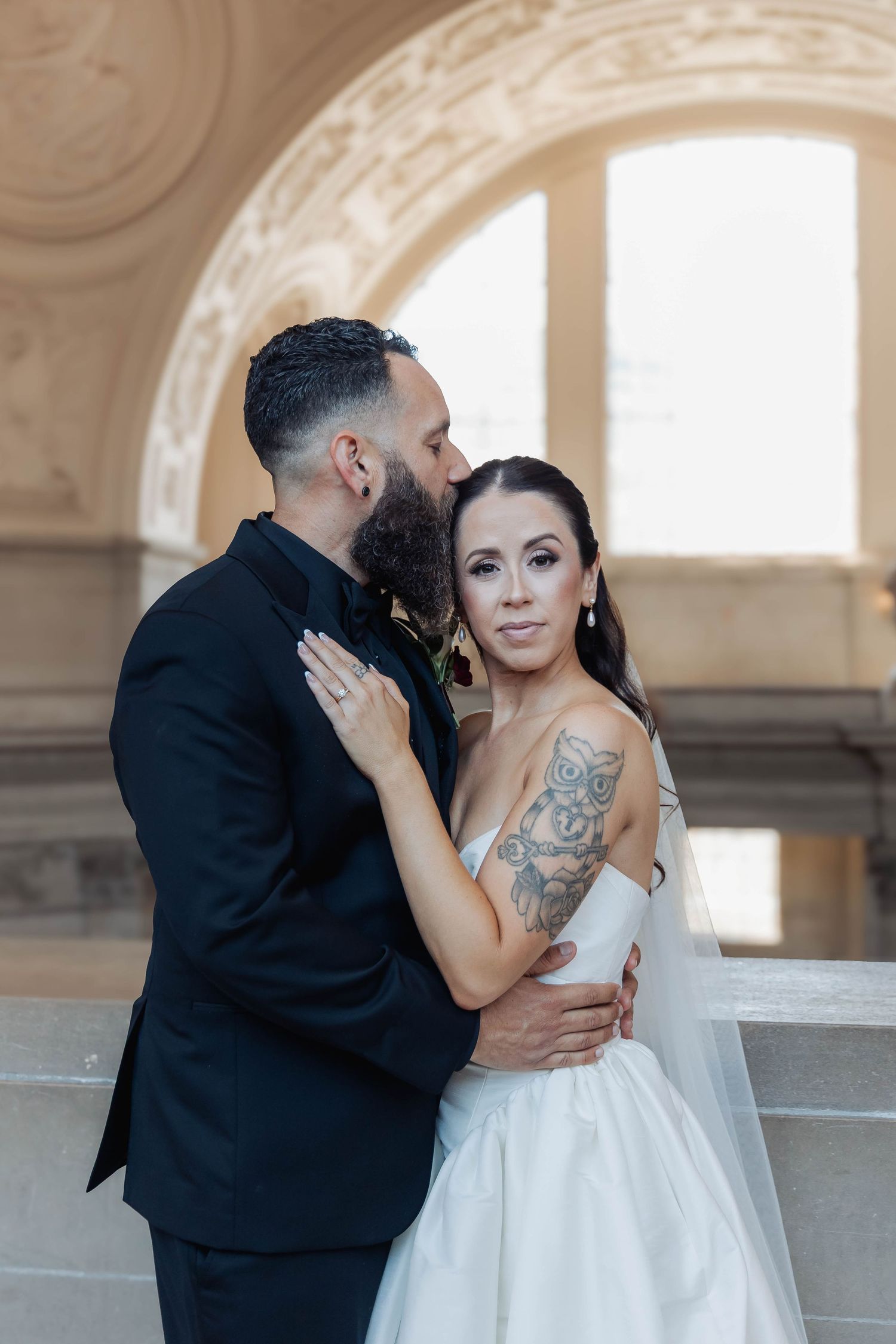 A groom in a black tuxedo kisses his tattooed bride in a white gown at an elegant wedding venue.