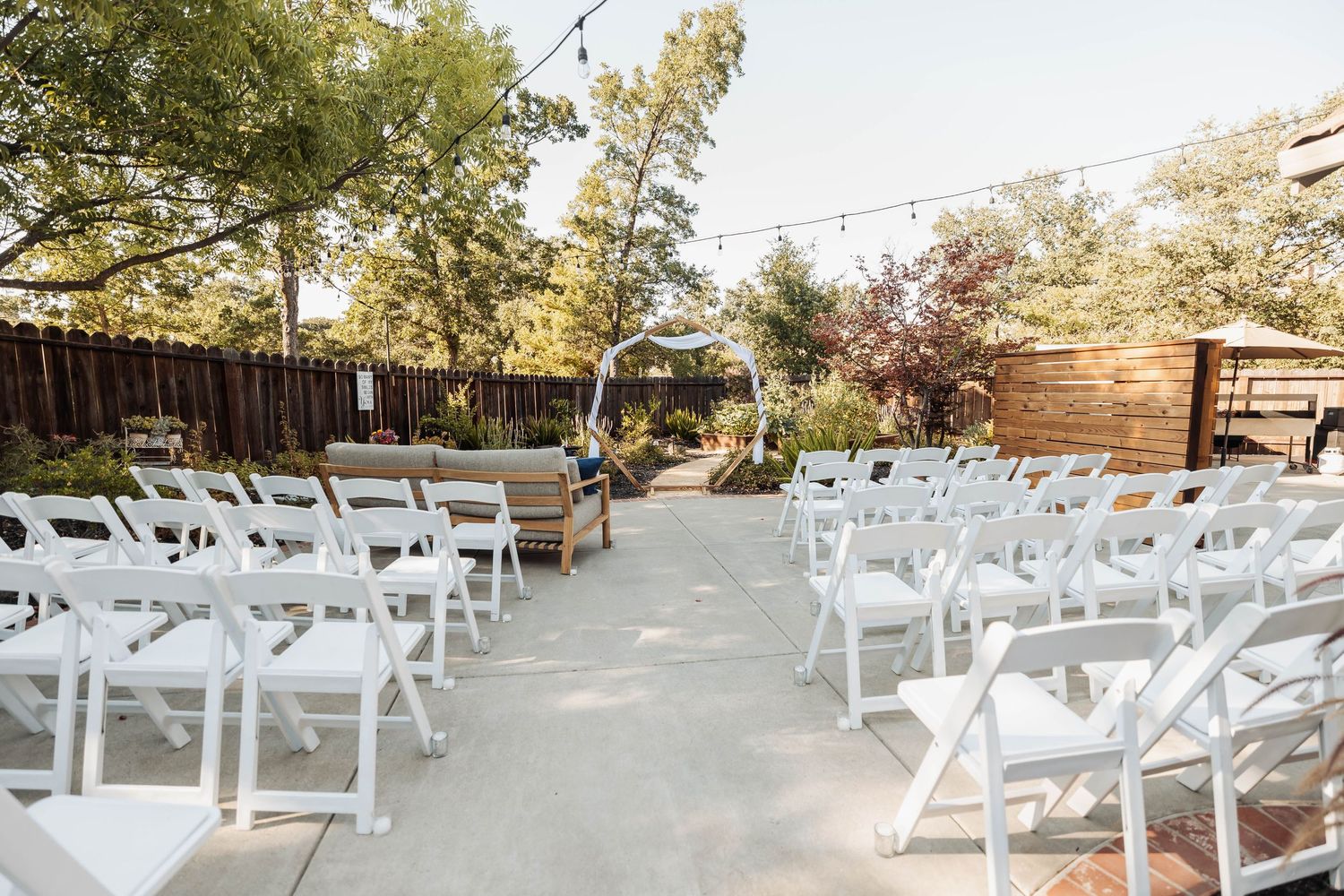 Outdoor wedding venue setup with white chairs and string lights under trees.