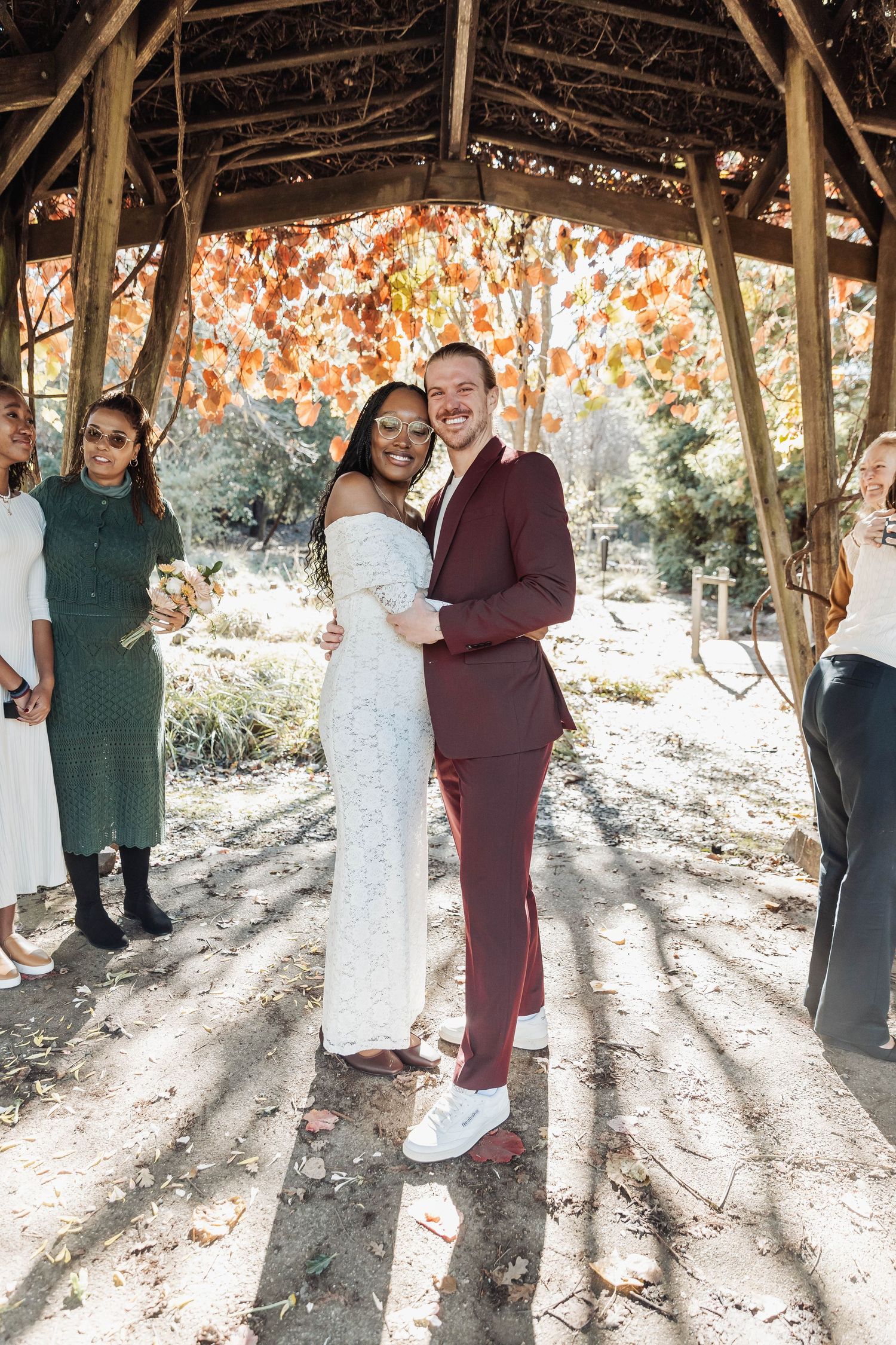 Wedding couple in white and burgundy sharing intimate moments under autumn trees.