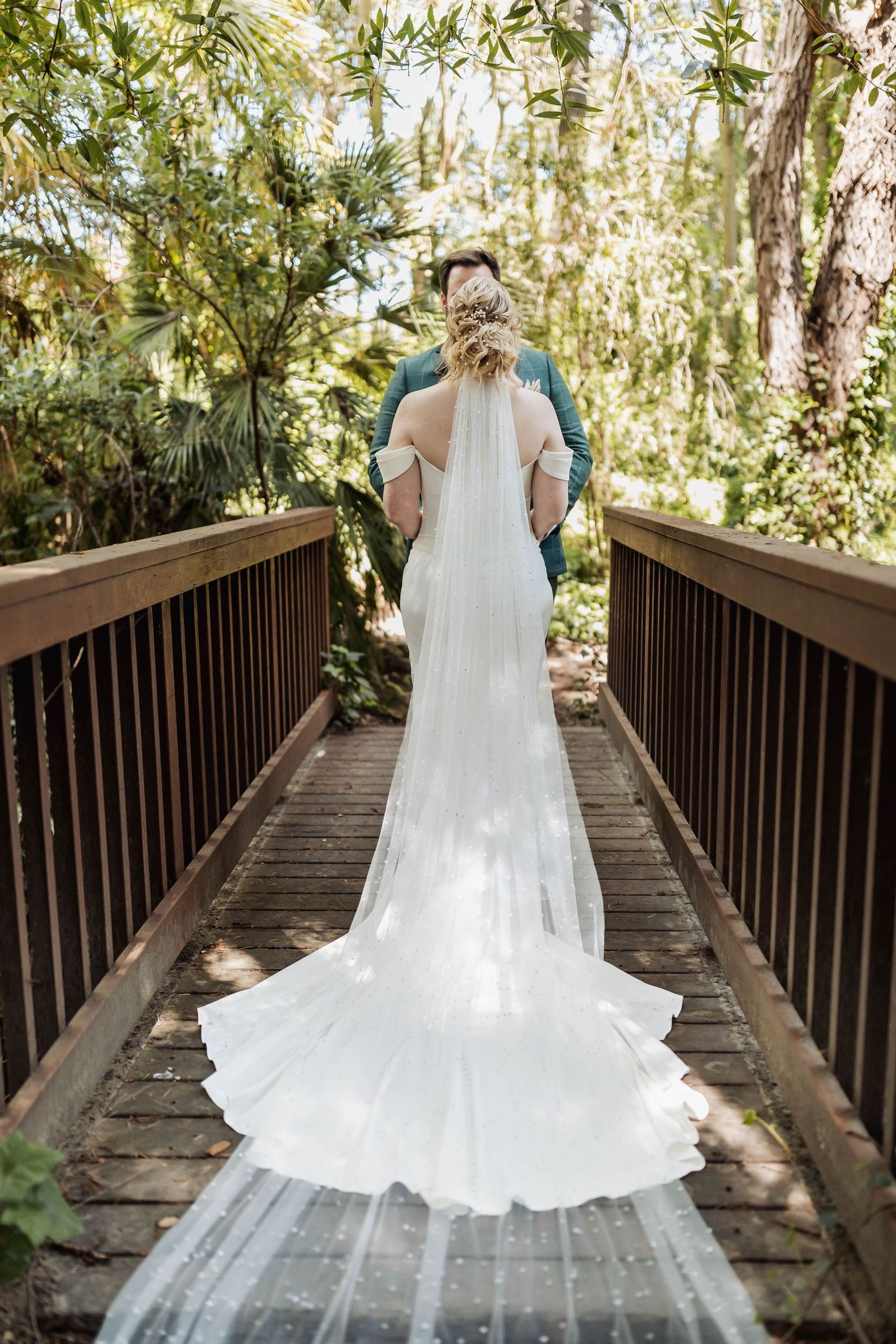 A flowing white wedding dress train cascades down a wooden bridge walkway surrounded by lush greenery.