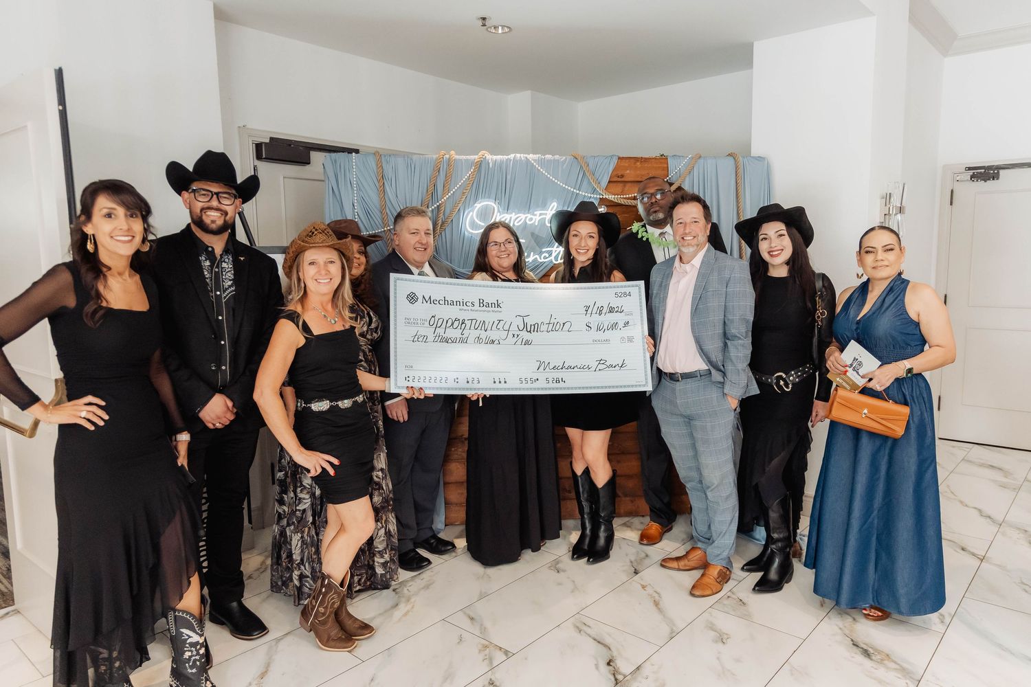 Group of elegantly dressed attendees pose with large charity check at Grit & Grace fundraising gala event.