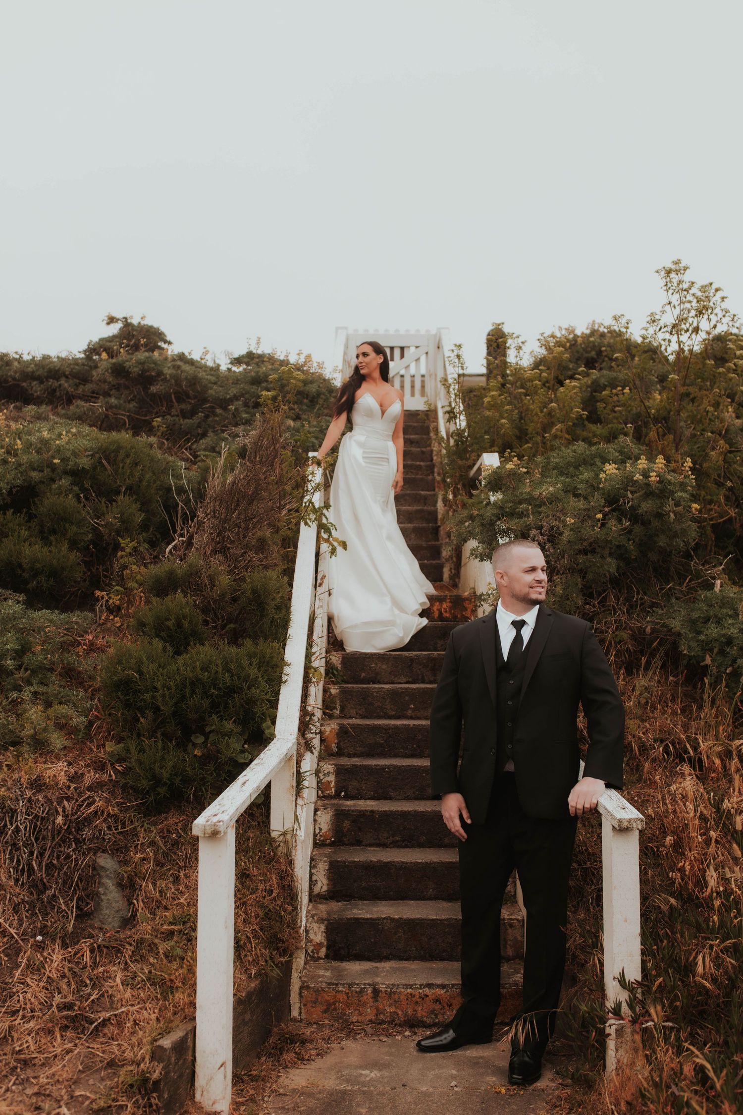 A bride and groom pose on rustic wooden stairs surrounded by coastal vegetation.