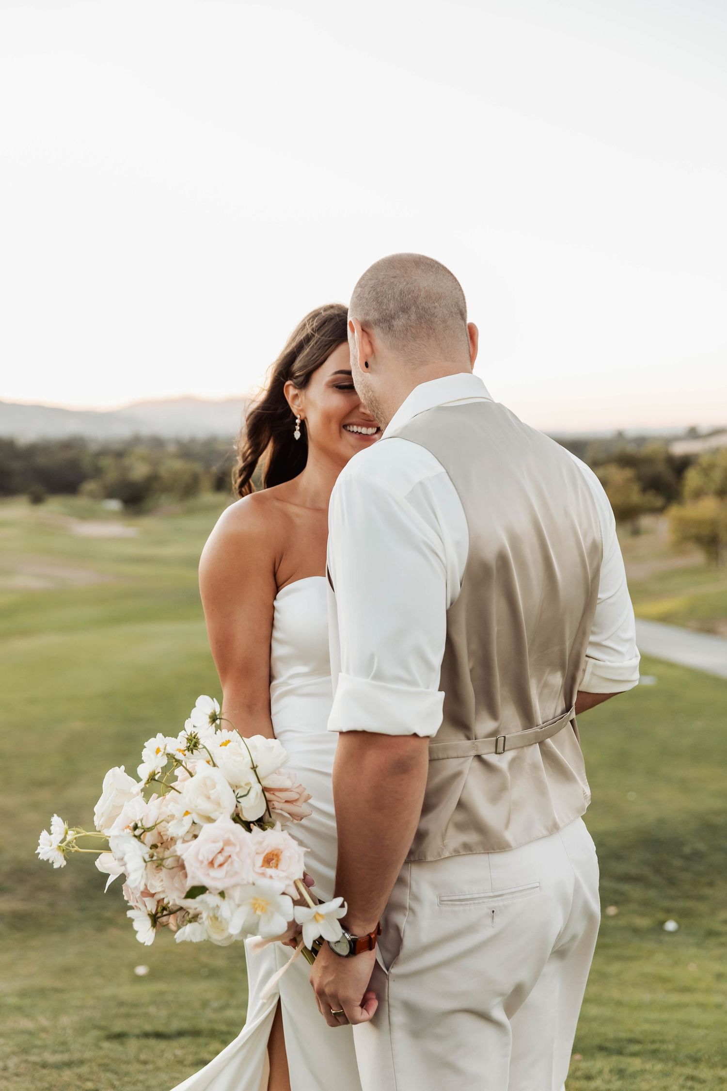 Couple in white and beige attire pose together on a golf course with white floral bouquet.