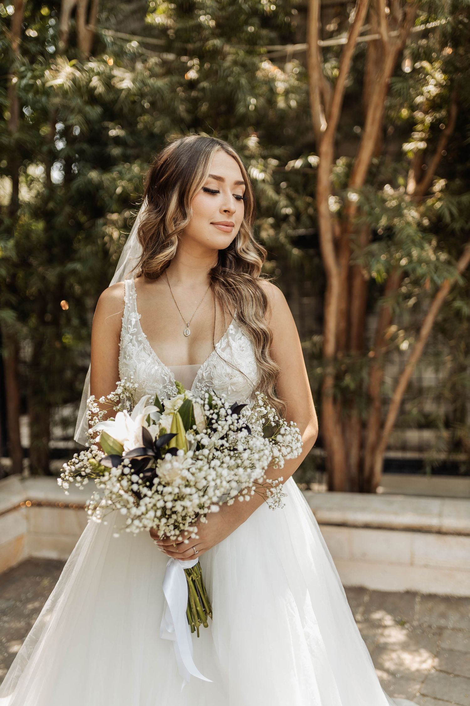 A bride in a white lace gown holds a delicate bouquet of white flowers and greenery in a sunlit outdoor setting.