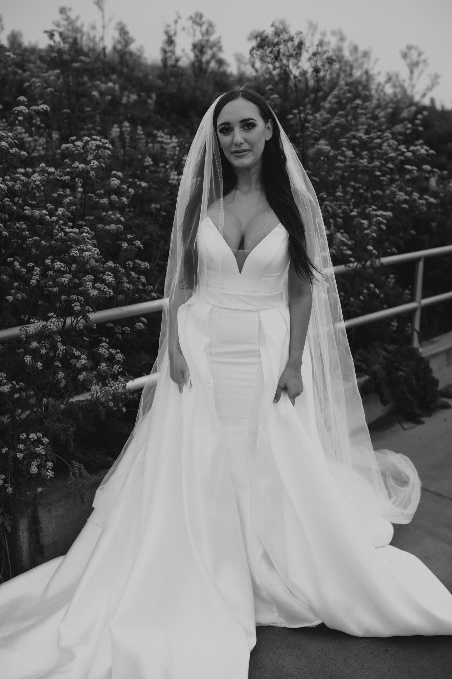 Elegant bride in fitted white wedding gown with flowing train and veil in garden setting.