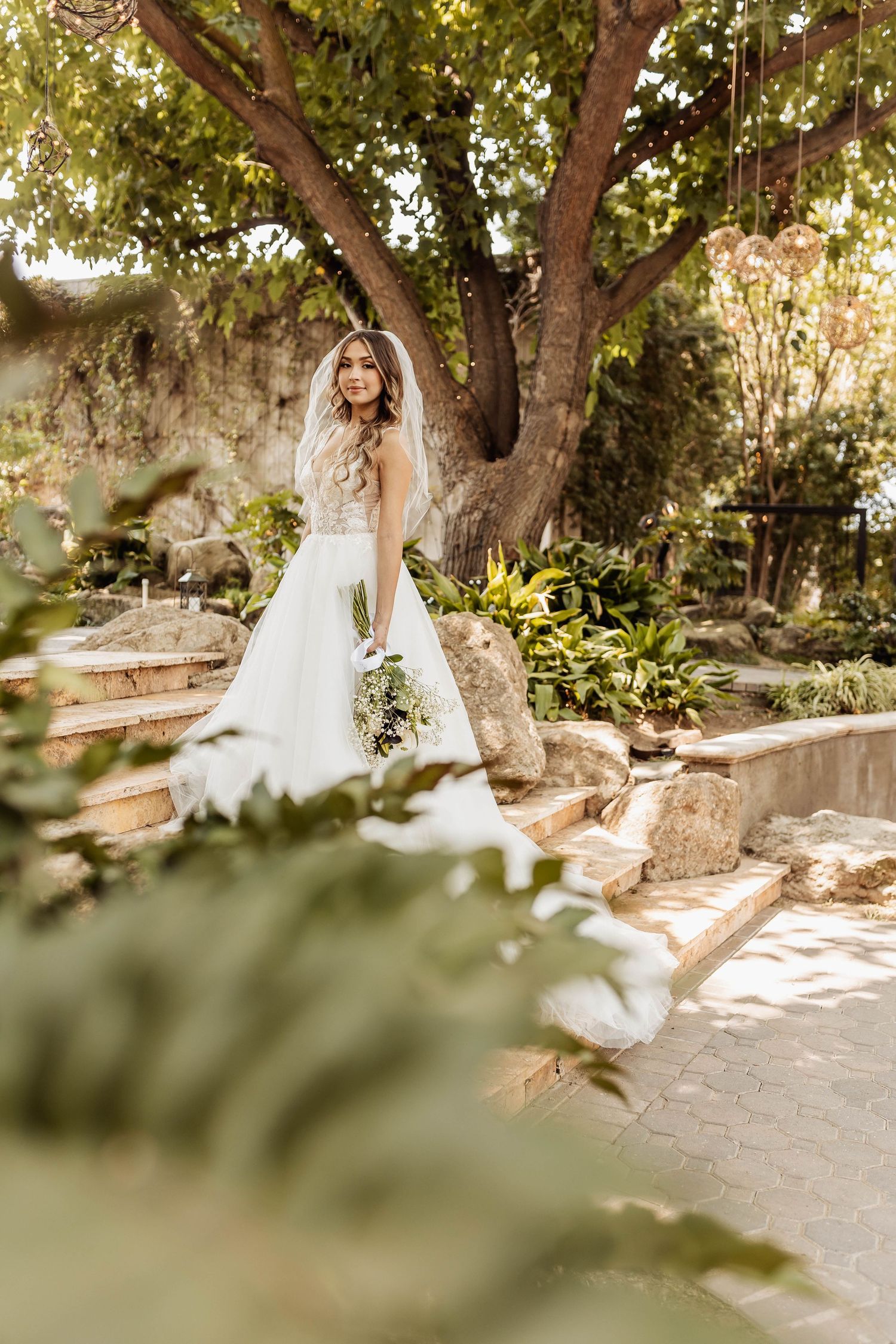 Romantic garden bridal portrait with flowing white dress against stone steps and lush foliage.