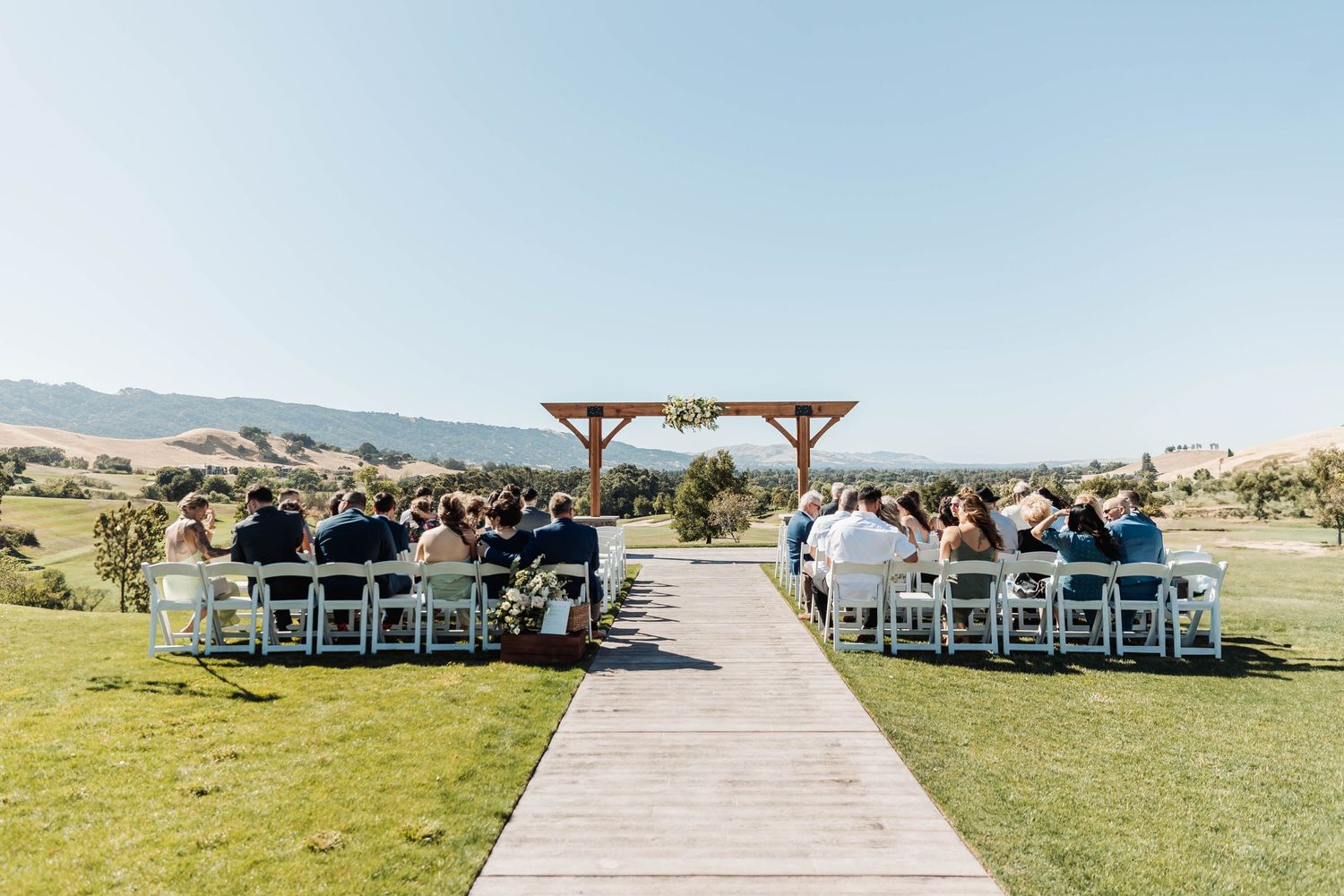 Outdoor wedding ceremony setup with wooden arch and white chairs on a grassy hill under blue skies.