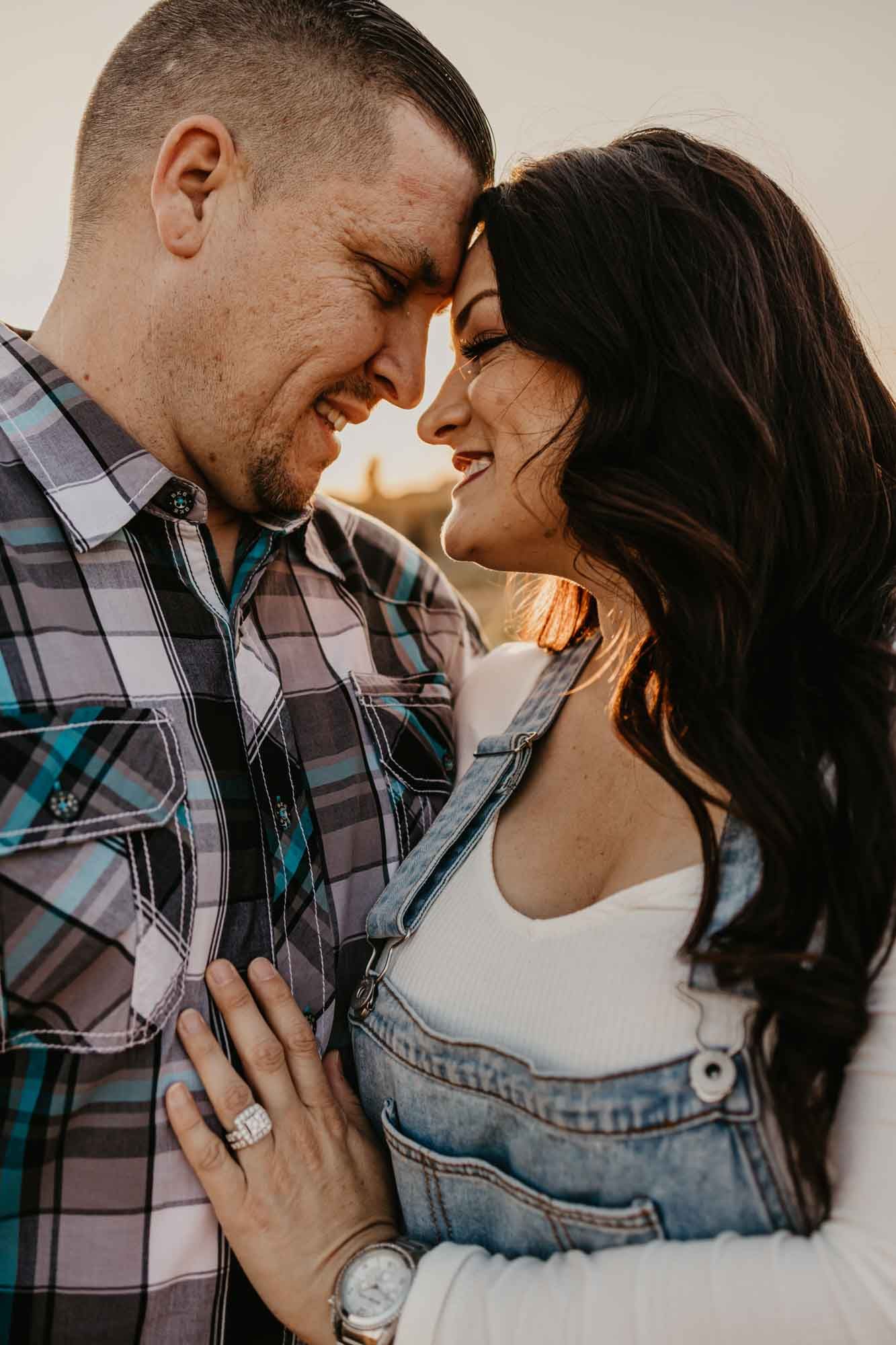 A romantic couple shares an intimate moment at sunset while wearing casual attire and showing off an engagement ring.