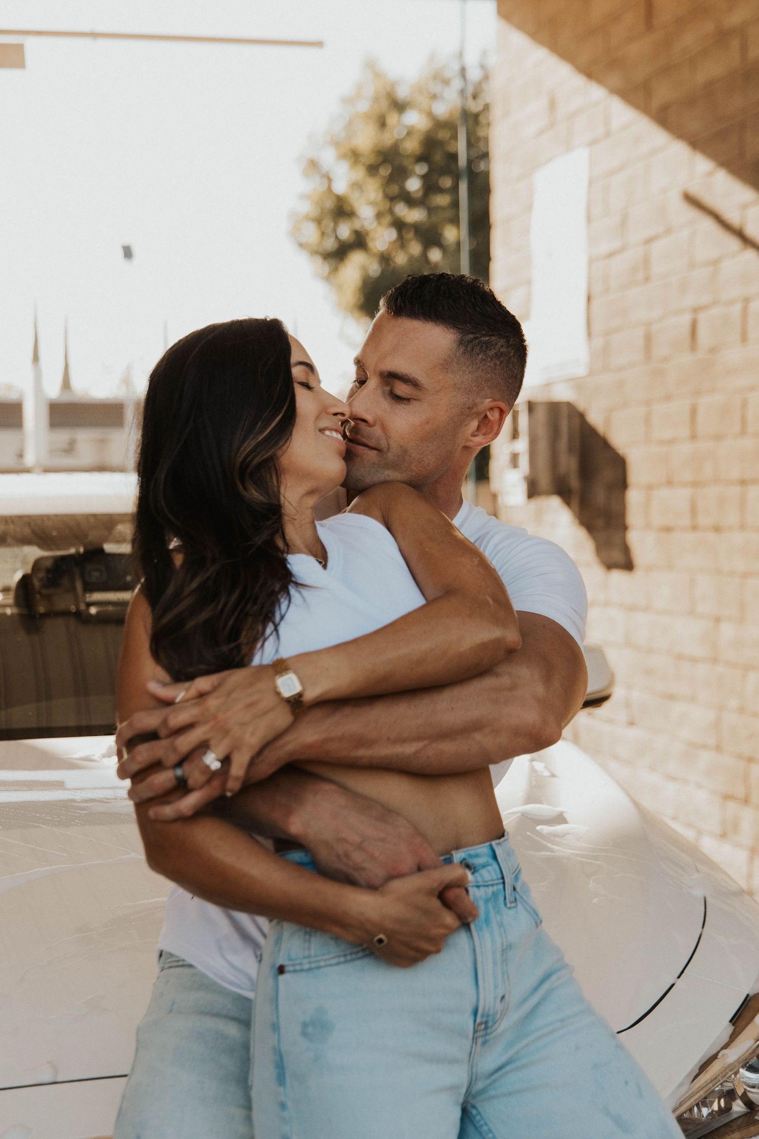 Couple in white tops and denim sharing romantic moments against brick wall.