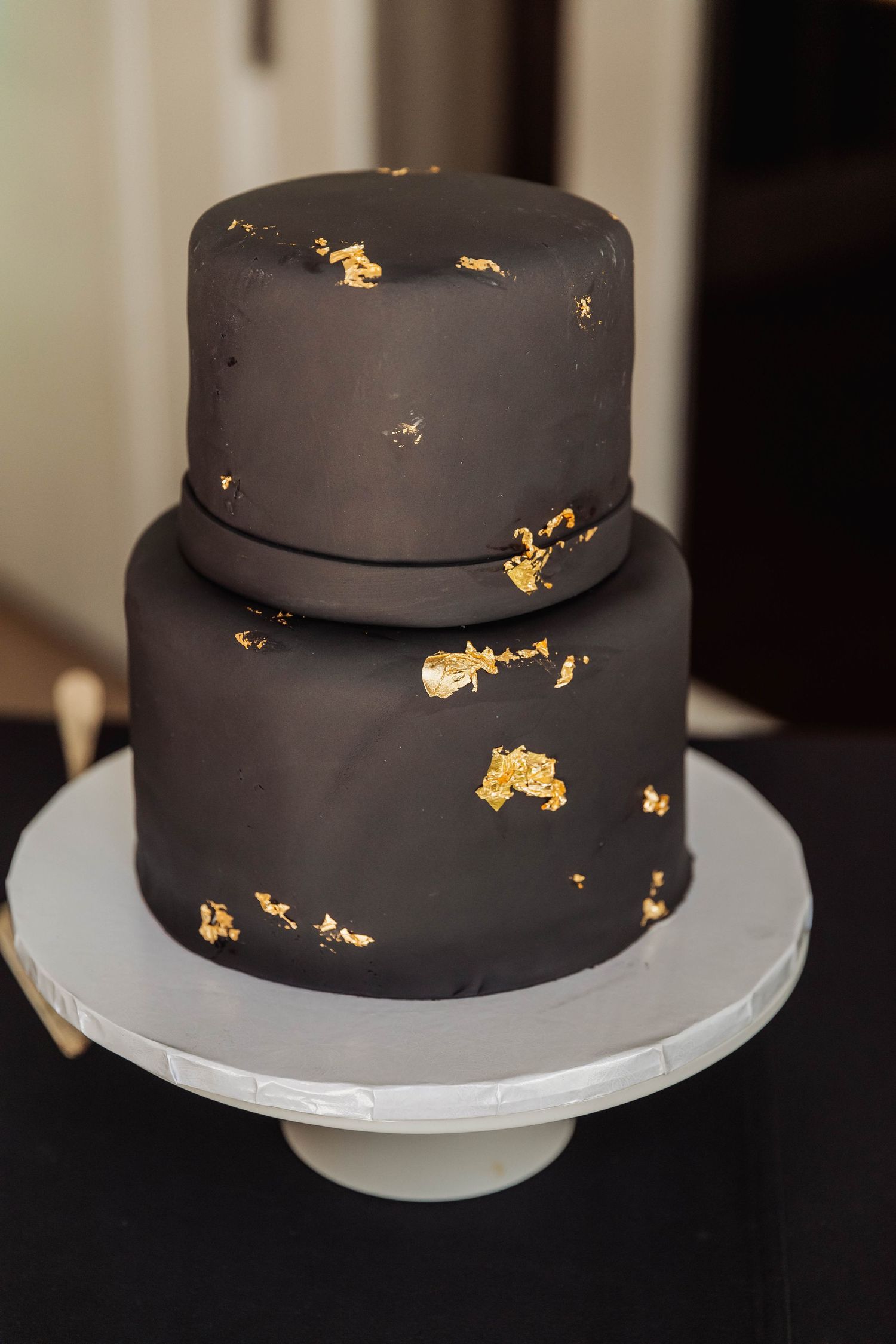 A modern black wedding cake with gold leaf accents displayed on a white cake stand.