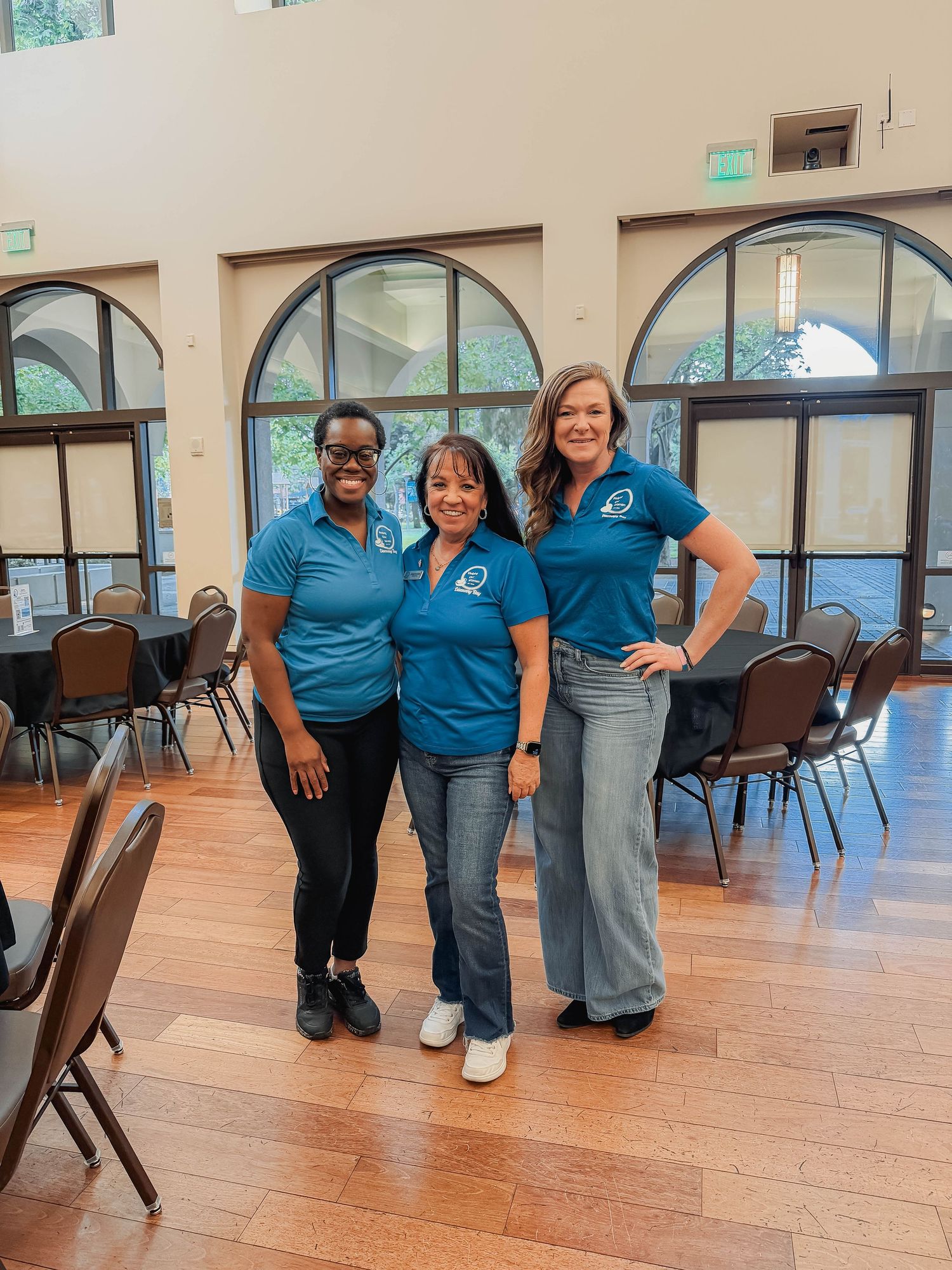 Three women in matching teal polo shirts posing together in an elegant event venue with arched windows.
