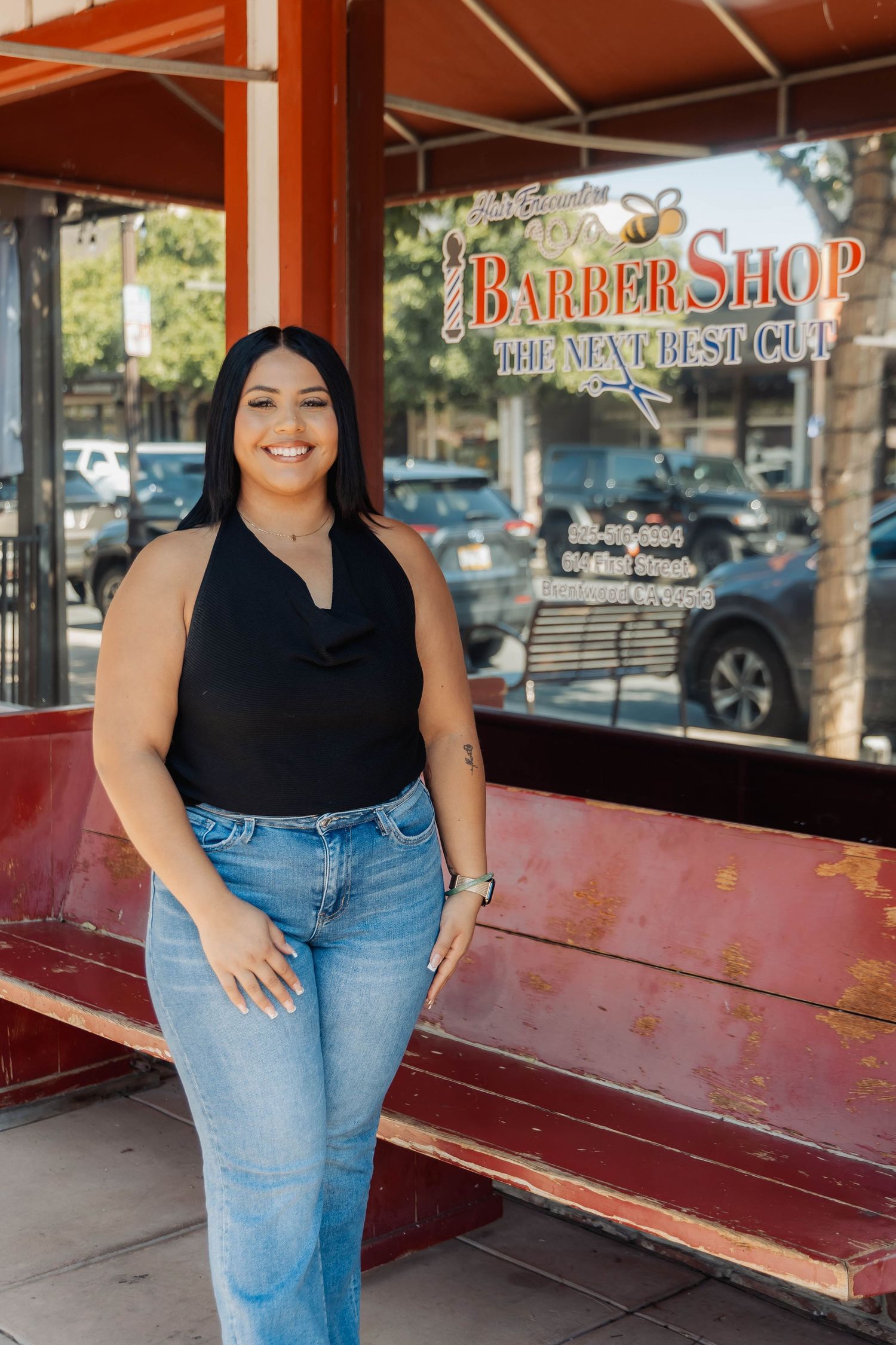Professional portrait of someone in a black top and jeans standing outside a barber shop storefront.