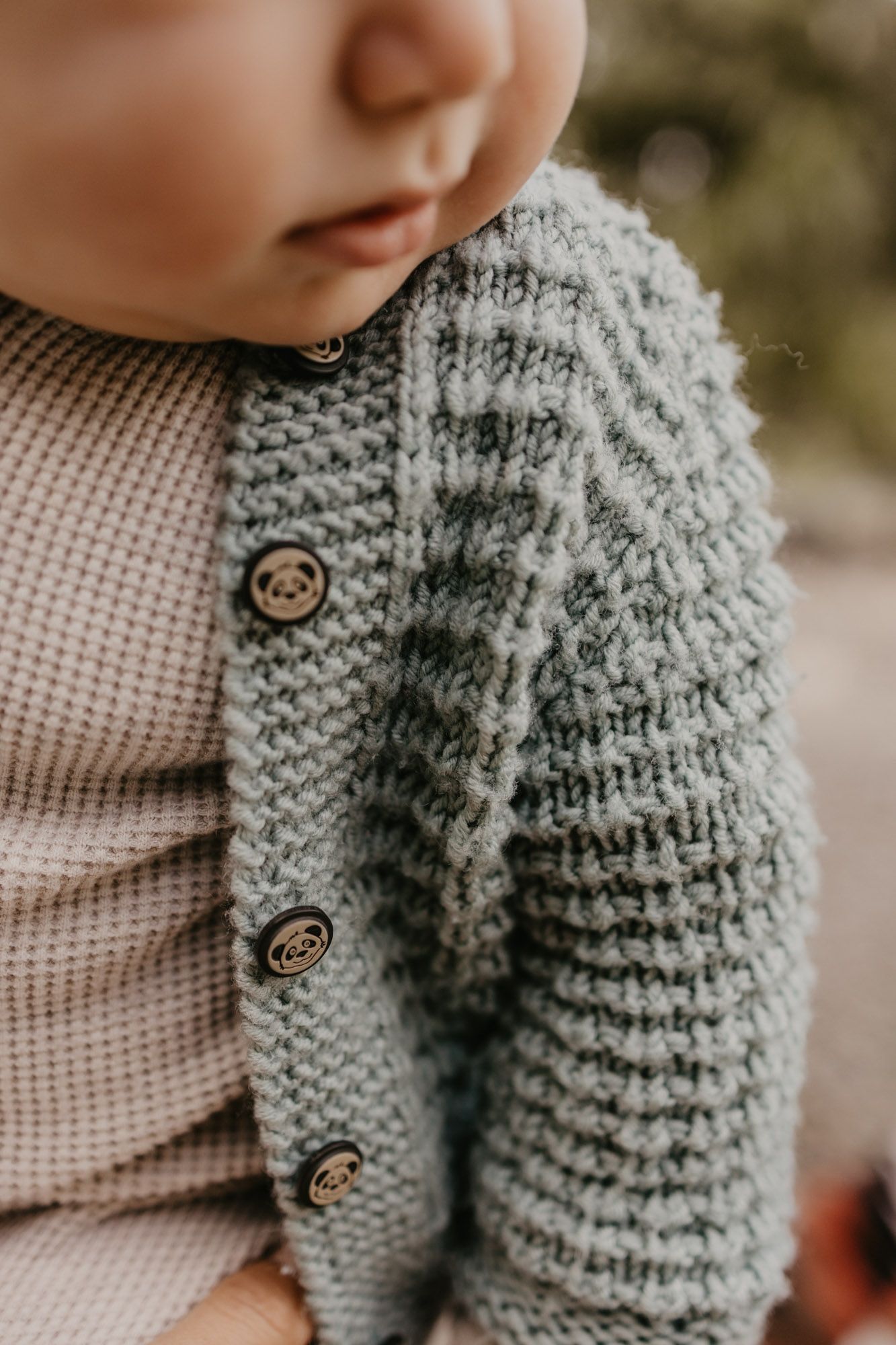 Detail shot of baby wearing cream-colored outfit with gray knitted cardigan with button details.