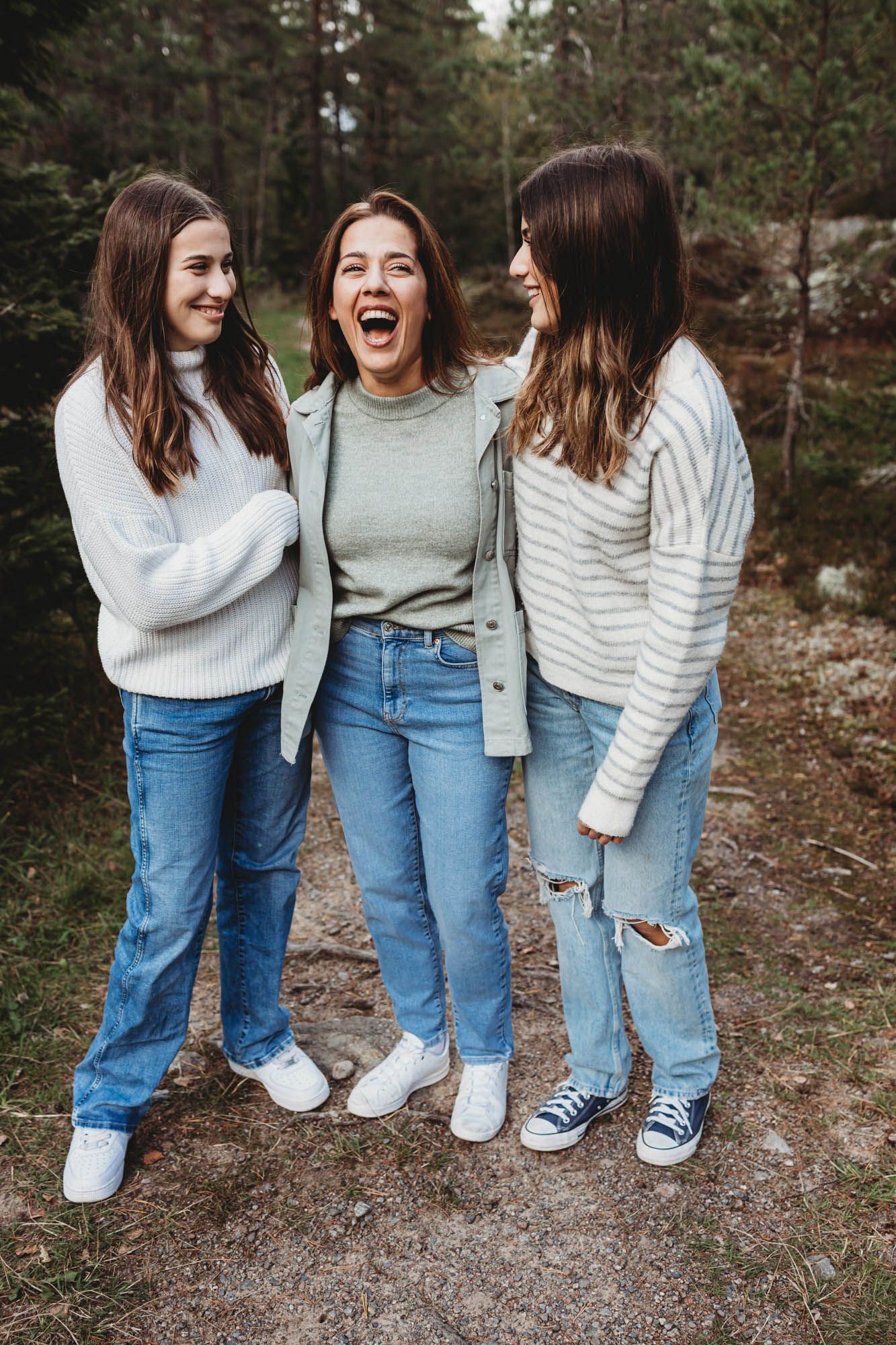 Three friends in sweaters and jeans share a hearty laugh on a hiking trail.