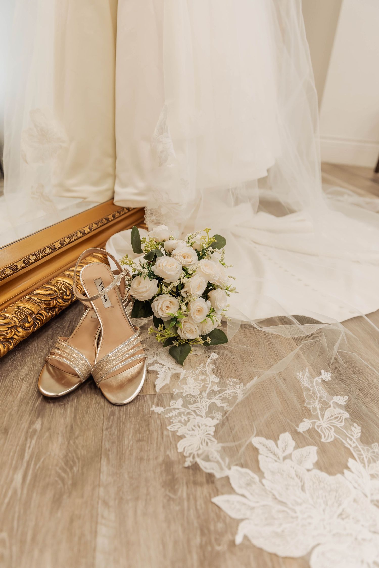 Wedding accessories including gold sandals, white bouquet and lace veil on wooden floor.